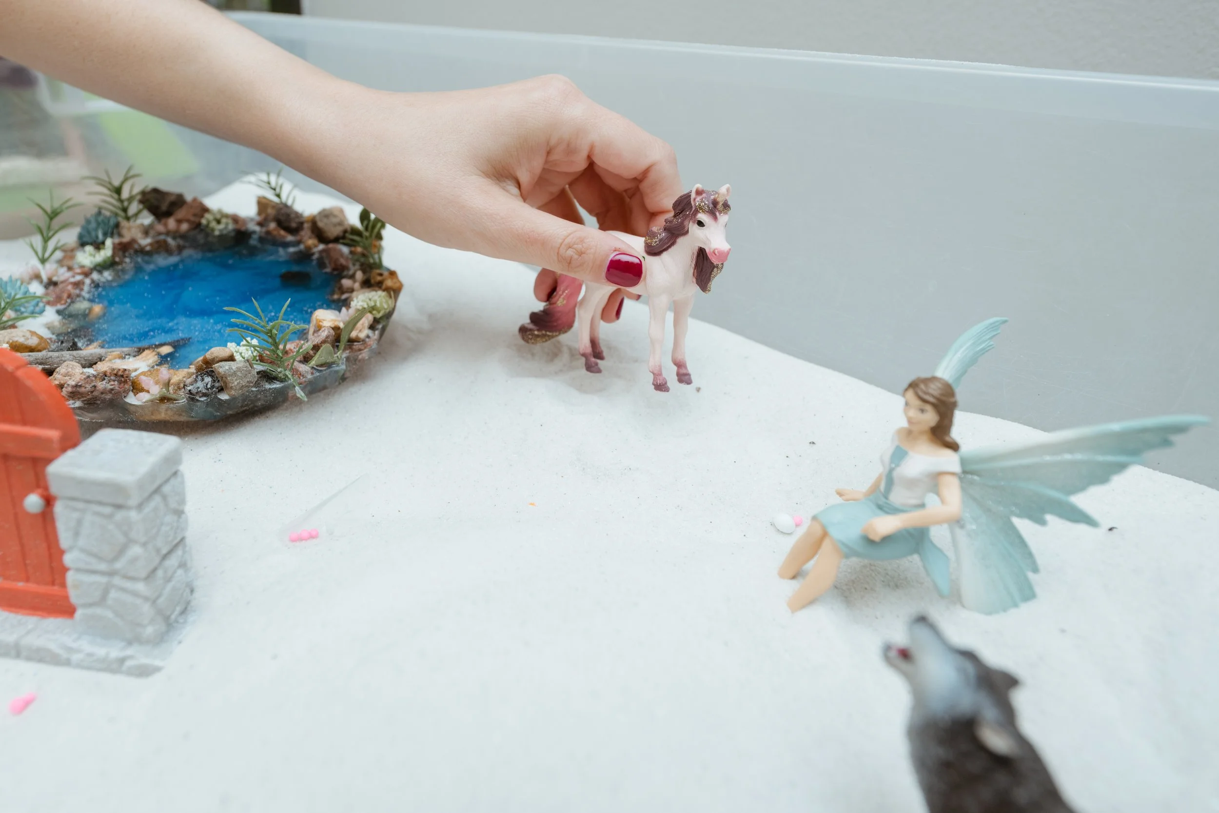 A person’s hand holding a green dragon figurine with wings, horns, and a necklace, sitting on white sand with small pink and white decorations, and a small white unicorn figurine in the background.