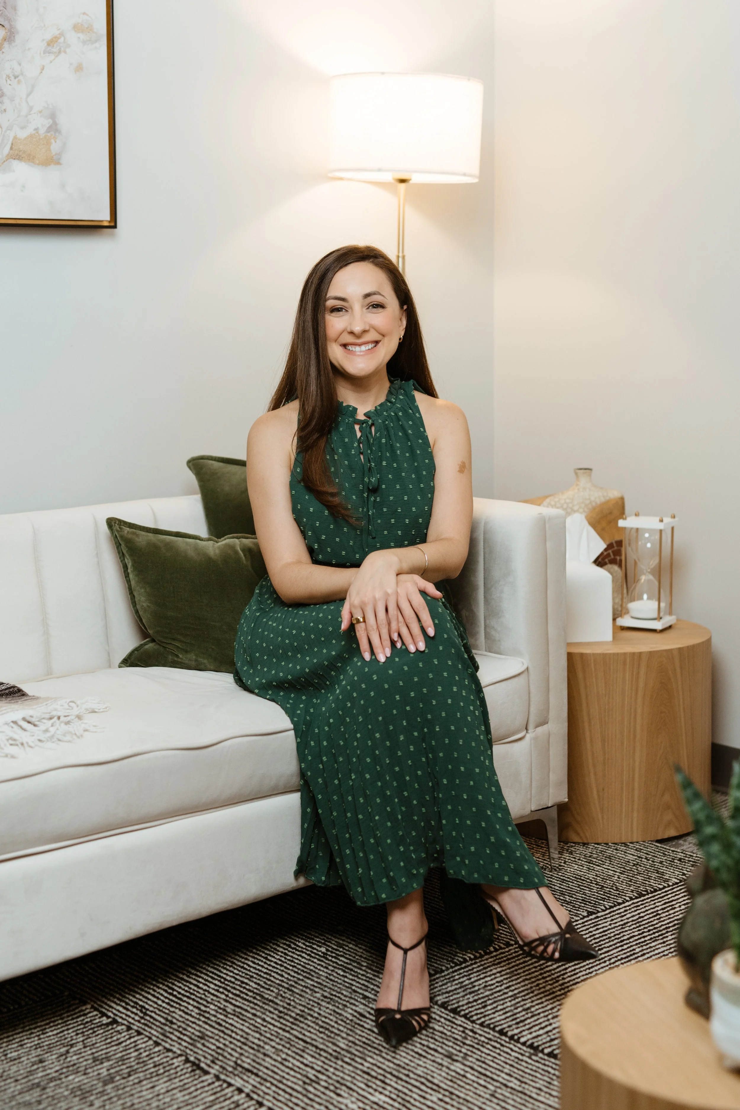 A smiling woman with long brown hair in a green polka dot dress standing in a room with a white couch, green pillow, and abstract artwork on the wall behind her.
