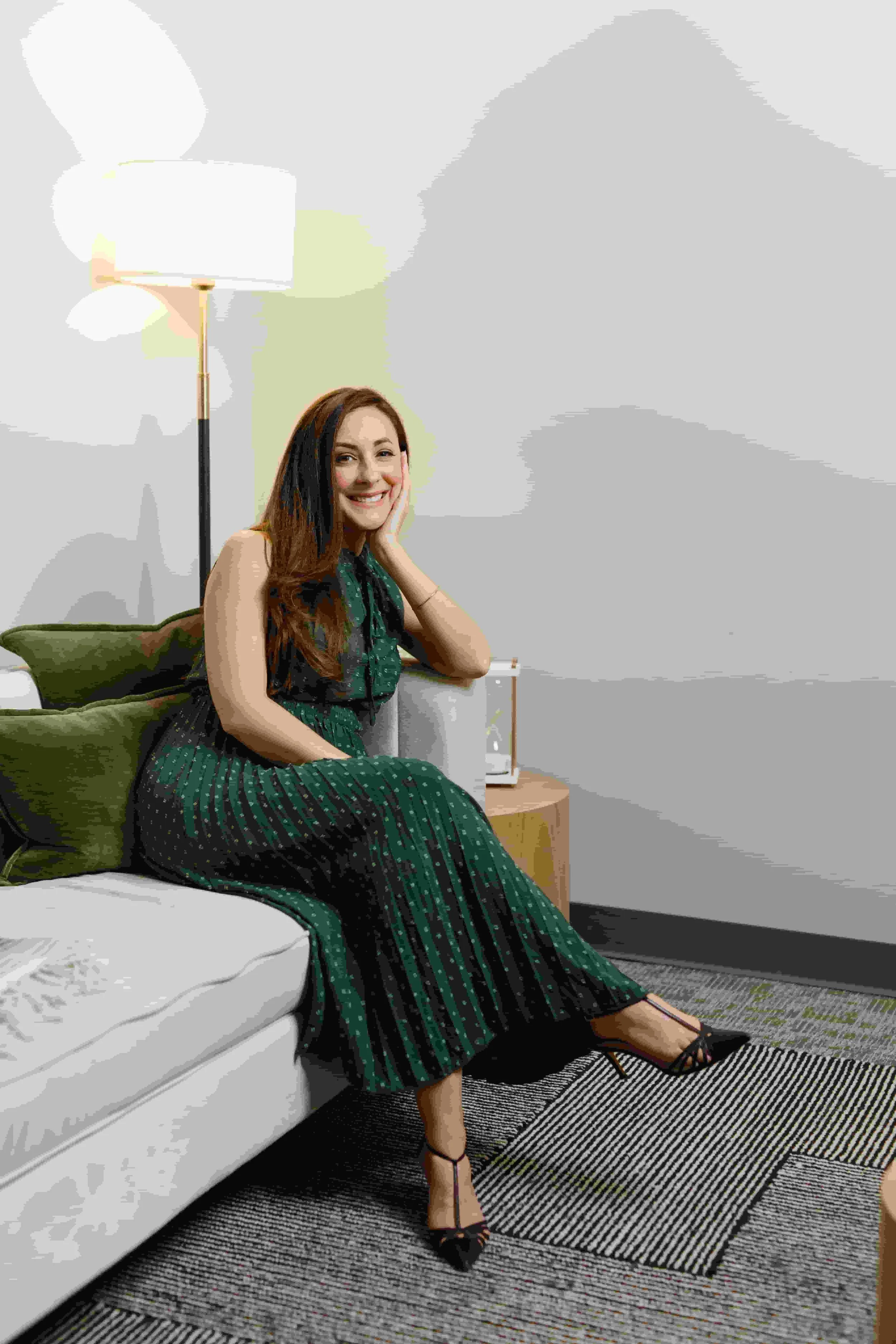 A smiling woman with long brown hair in a green polka dot dress standing in a room with a white couch, green pillow, and abstract artwork on the wall behind her.