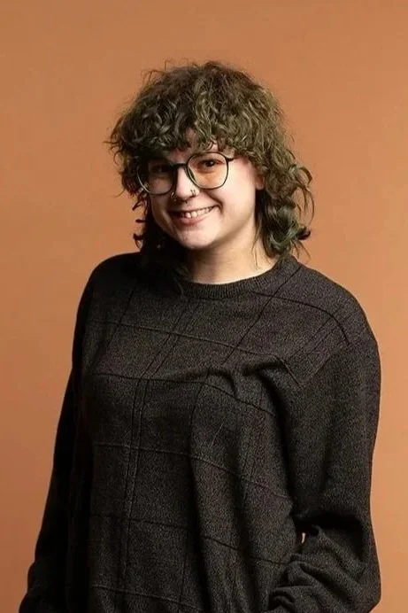 A young woman with curly hair and glasses smiling, wearing a dark patterned sweater, standing against a plain brown background.