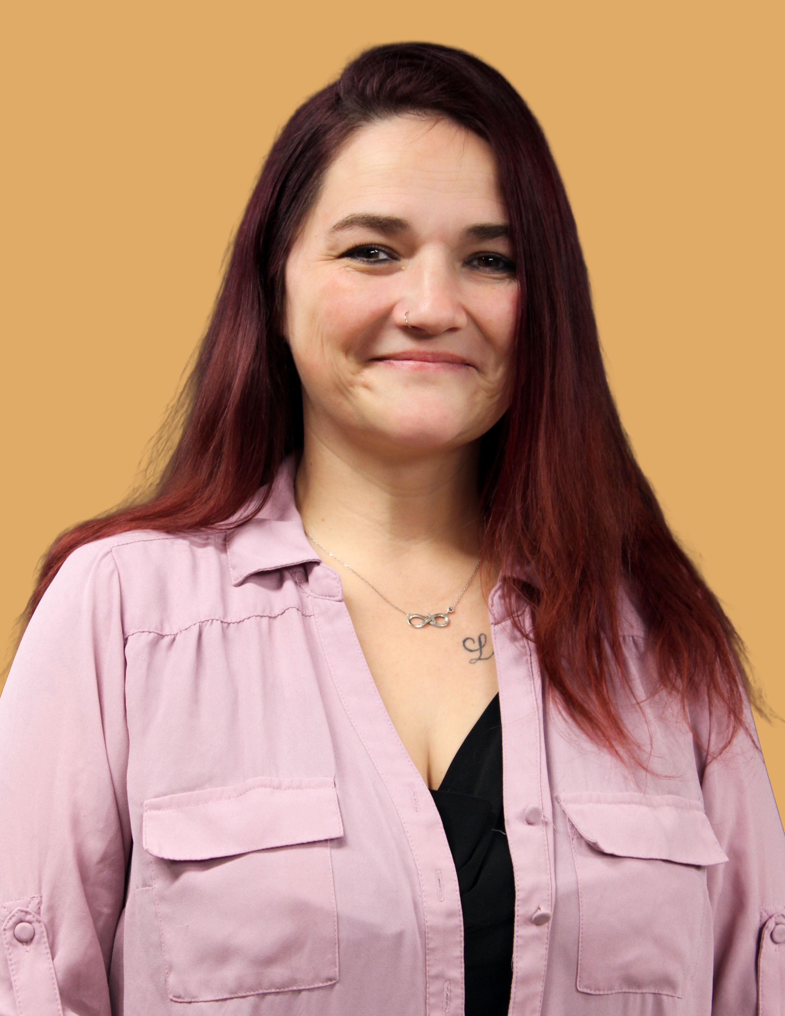 A woman with long, dark red hair, wearing a pink button-up shirt, a black top, a silver infinity necklace, and a nose ring, standing against a yellow background.