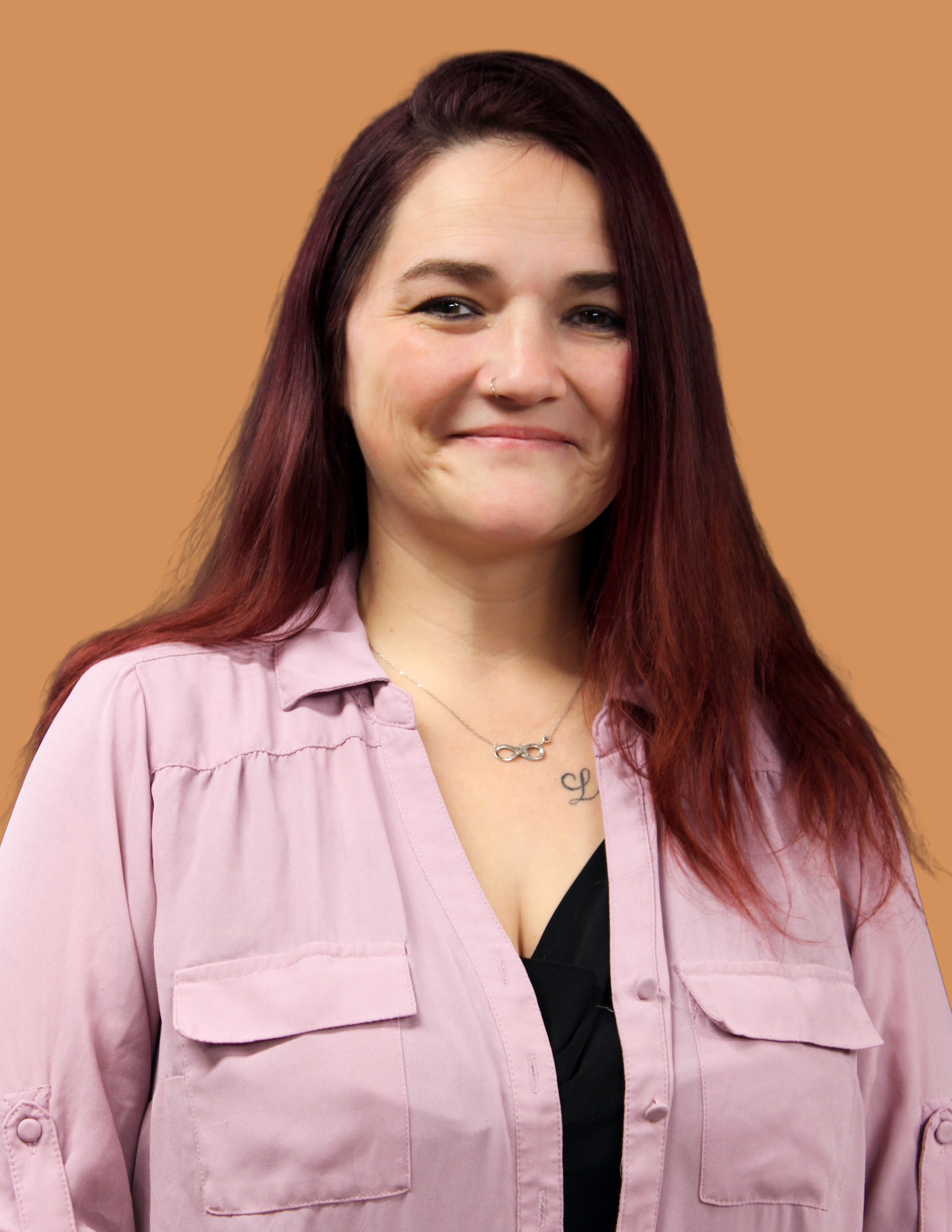 A woman with shoulder-length red hair, wearing a pink button-up shirt, a black top underneath, a silver infinity necklace, and a nose piercing, smiling against a tan background.