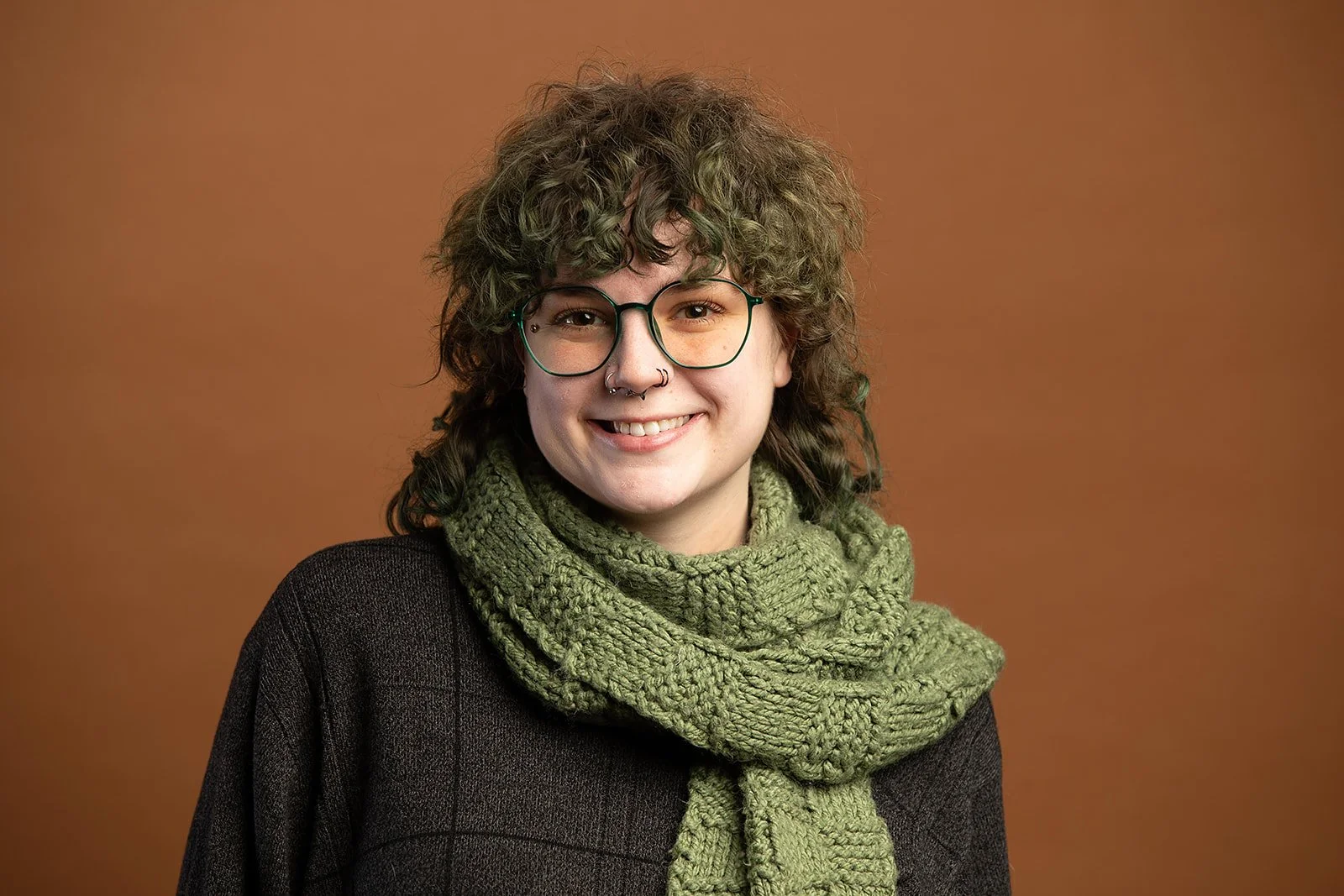 Young woman with curly hair, glasses, and septum piercing, smiling, wearing a green knitted scarf and a dark top, standing against a plain brown background.