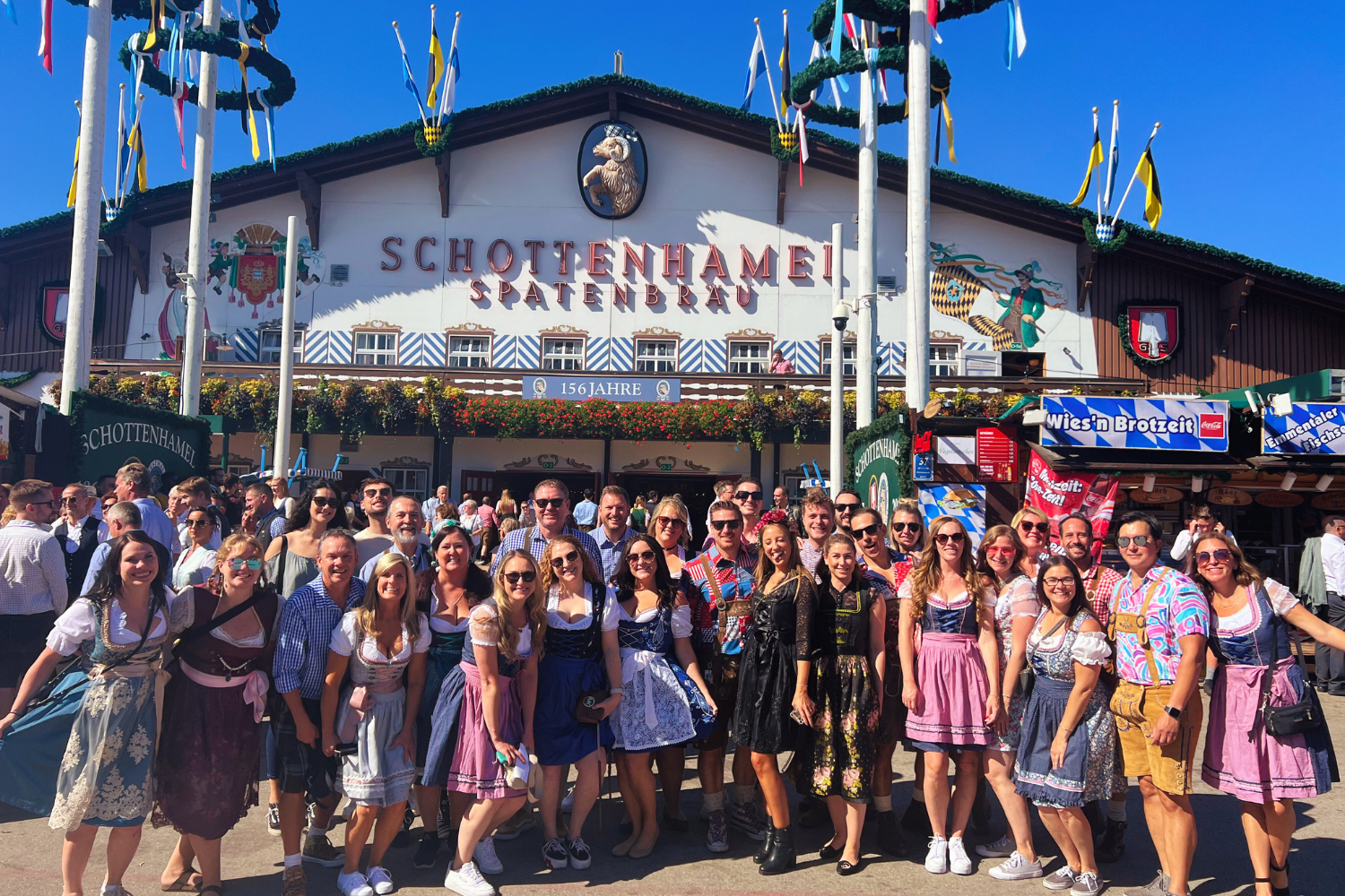 A group of people wearing traditional lederhosen and dirndls celebrating at Oktoberfest in Germany, smiling and raising steins of beer in a lively festival tent.