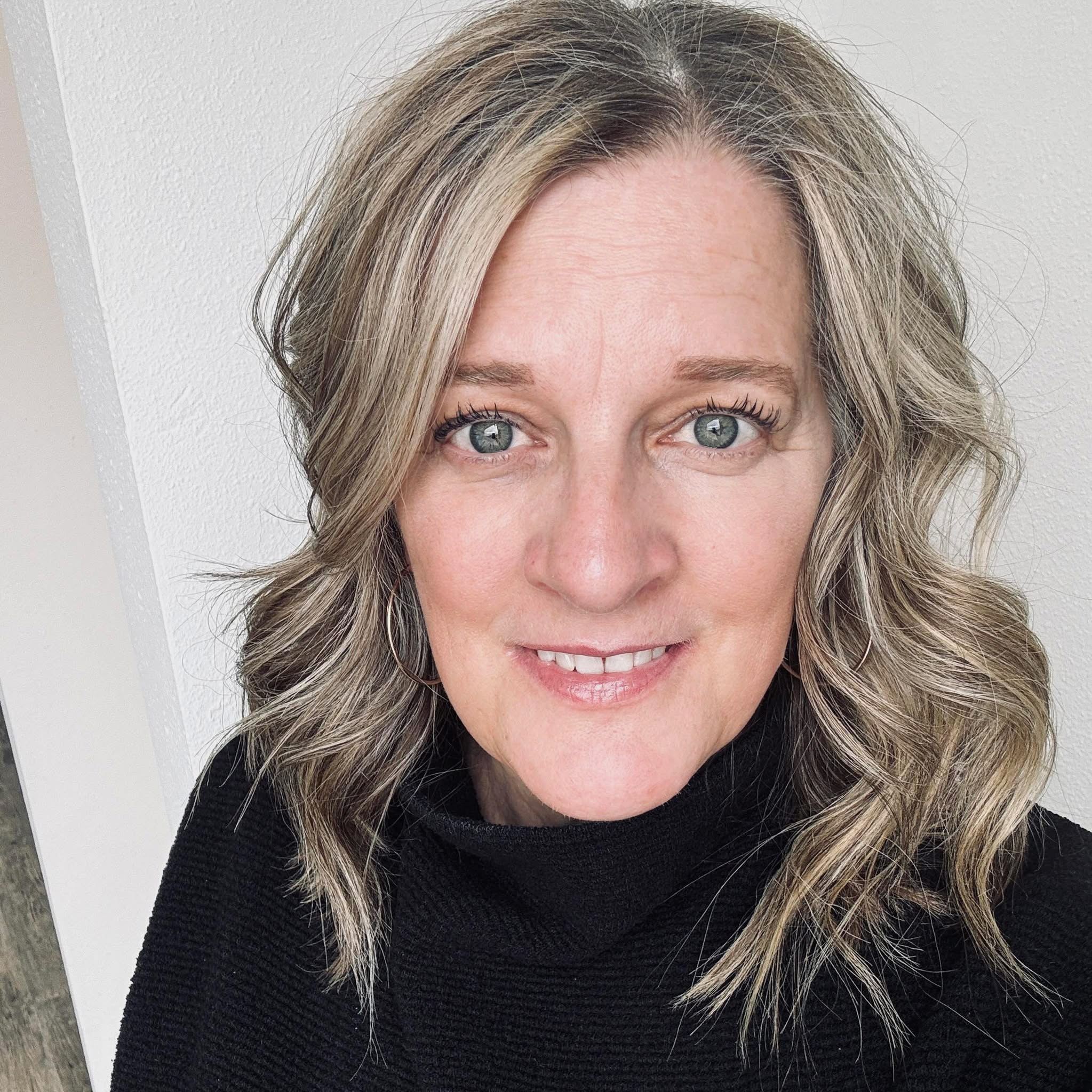 Close-up image of a woman with blonde, wavy hair and blue eyes, smiling slightly, wearing a black top, against a white background.