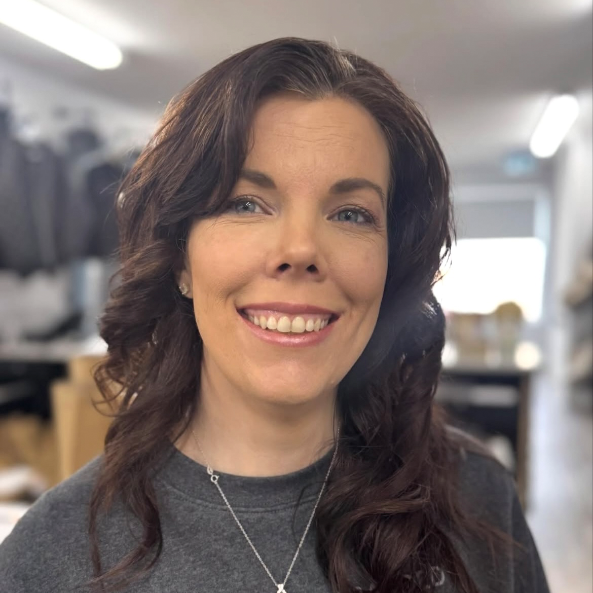 A woman with wavy dark brown hair and light eye color smiling in an indoor setting.
