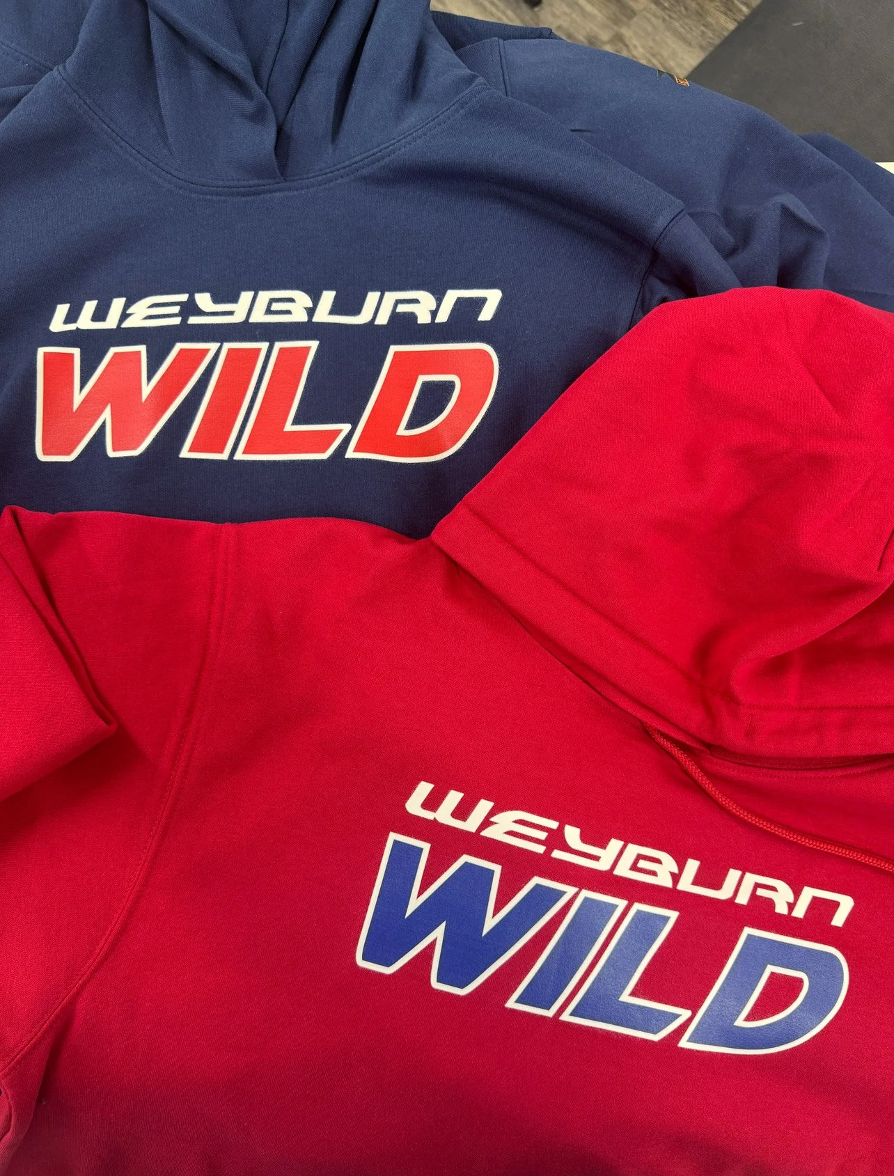 Close-up of two sweatshirts, one blue and one red, both with 'WILD' written on the front in large capital letters and 'WAYBURN' written above it. The sweatshirts are lying on a flat surface.