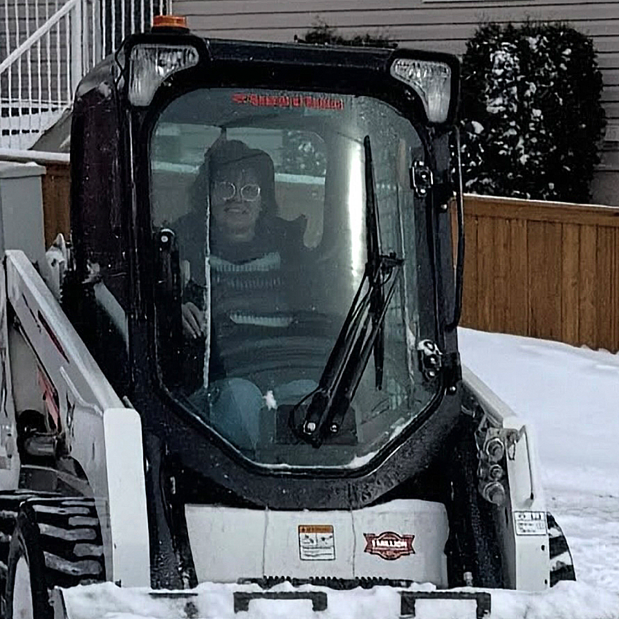 Person wearing glasses inside a snow-covered skid-steer loader with a the back of a house and fence in the background.