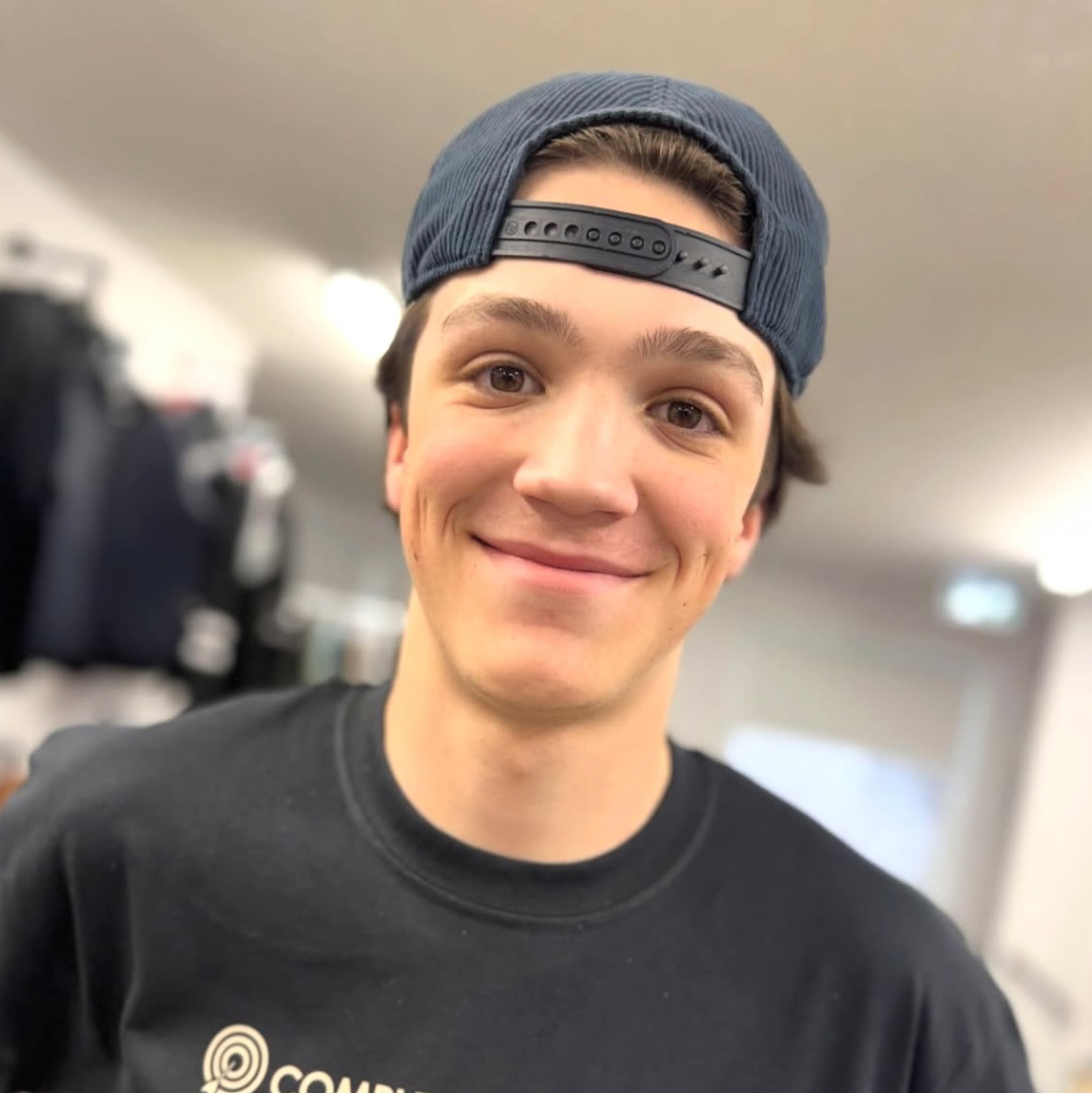 A young man smiling, wearing a backward baseball cap and a black t-shirt, in an indoor setting.