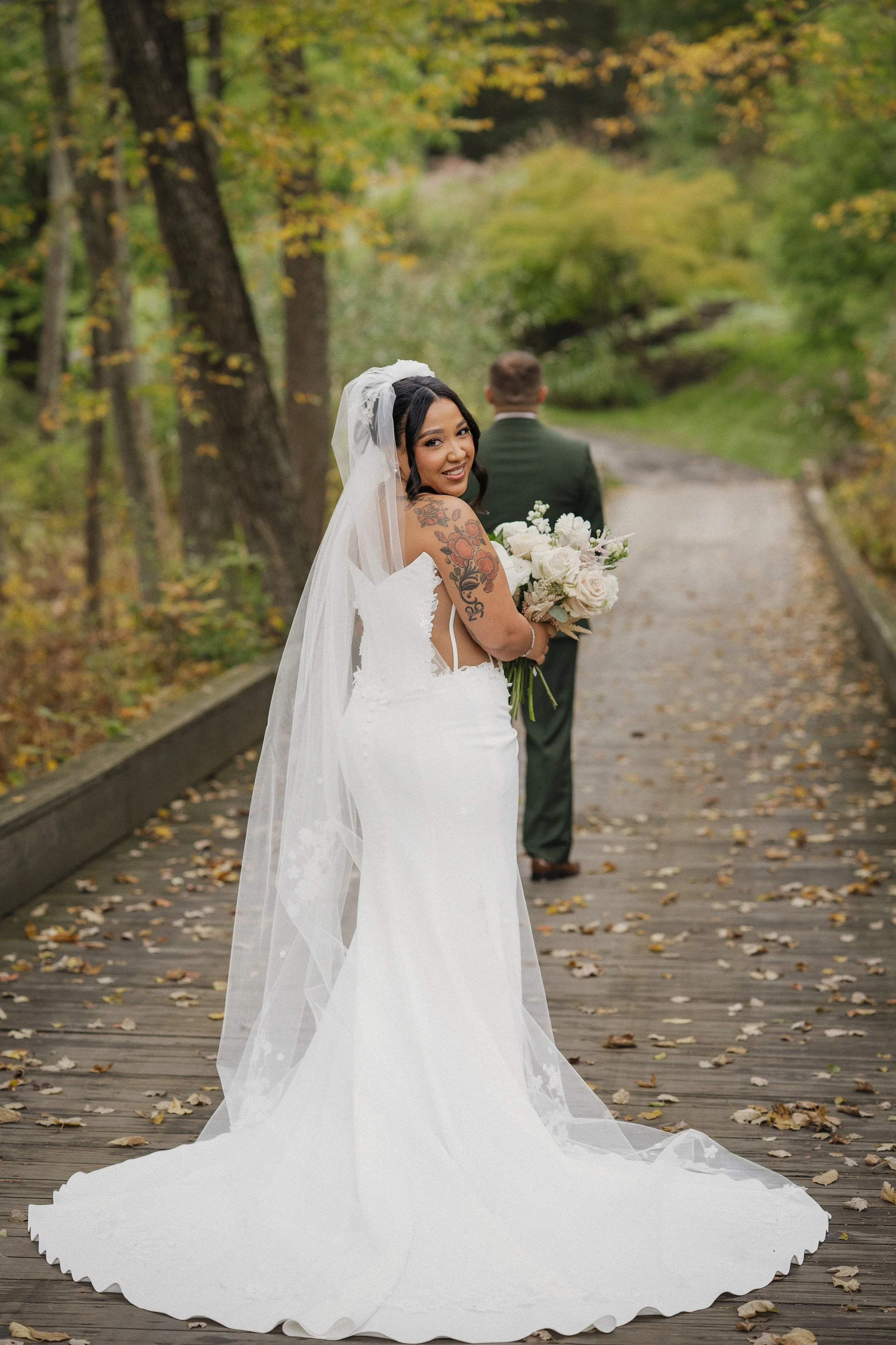 A bride in a white wedding dress holding a bouquet, smiling and turning back, with a groom in a green suit in the background on a wooden path surrounded by autumn trees.