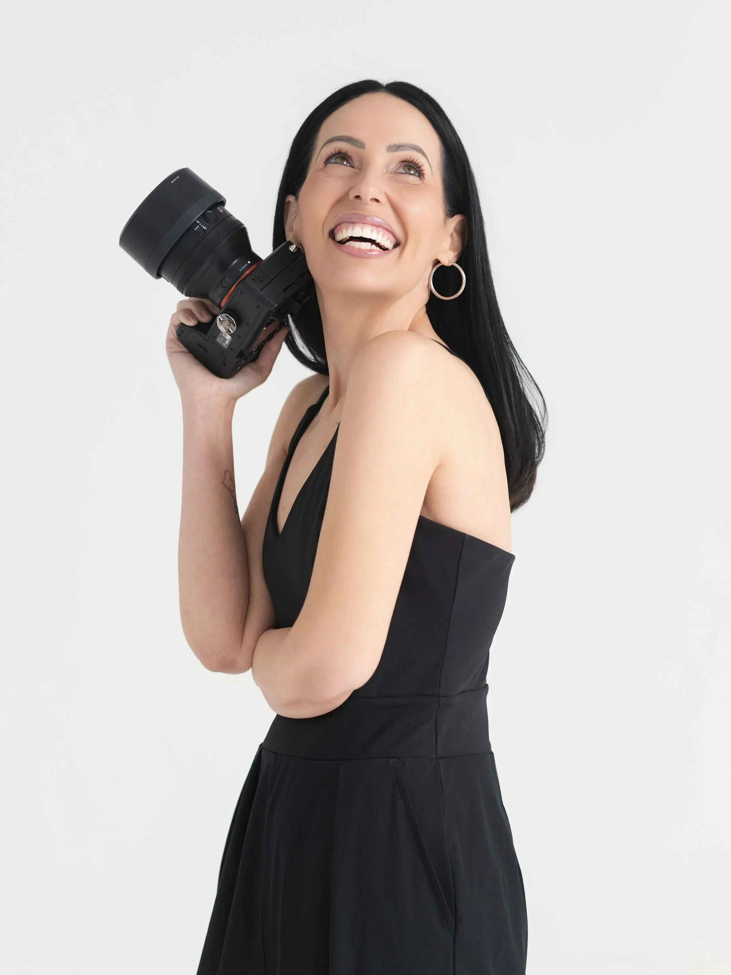 A woman with black hair, hoop earrings, wearing a black dress, smiling and holding a professional camera to her ear against a white background.