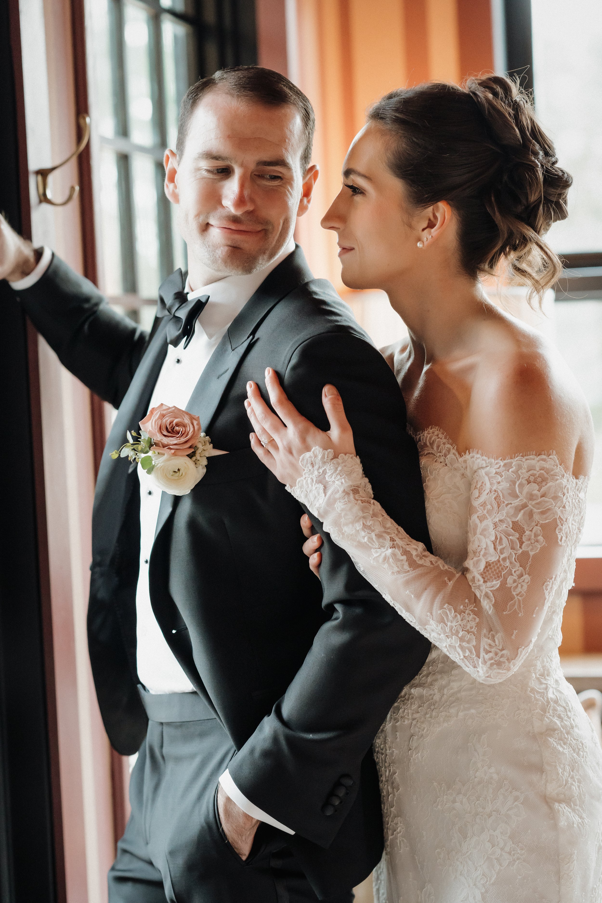 A bride and groom dressed in wedding attire, sharing a tender moment indoors near a window.