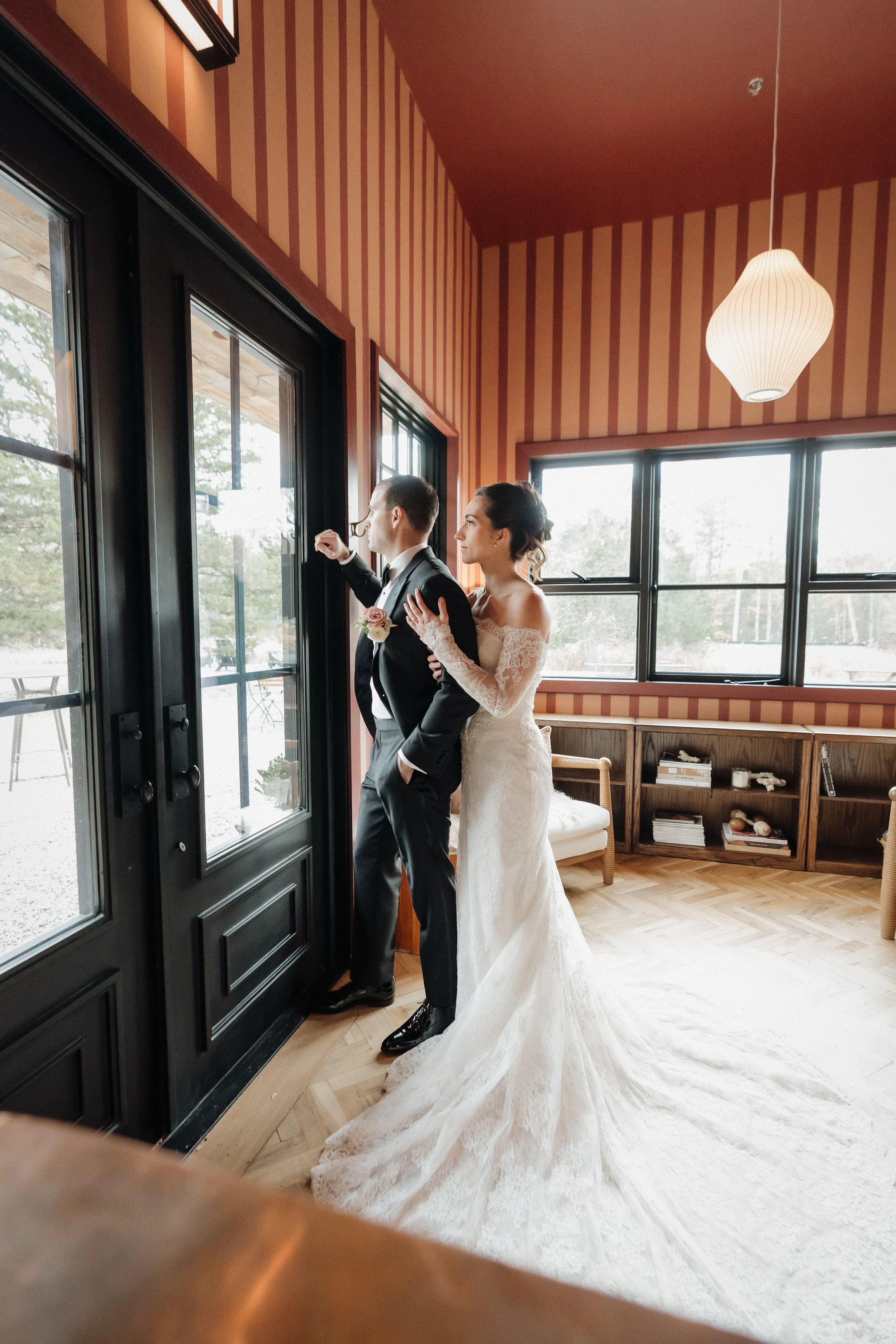 A bride and groom standing indoors by glass doors, with the bride in a white lace wedding dress and the groom in a black suit. The groom looks out the window, while the bride lovingly looks at him.