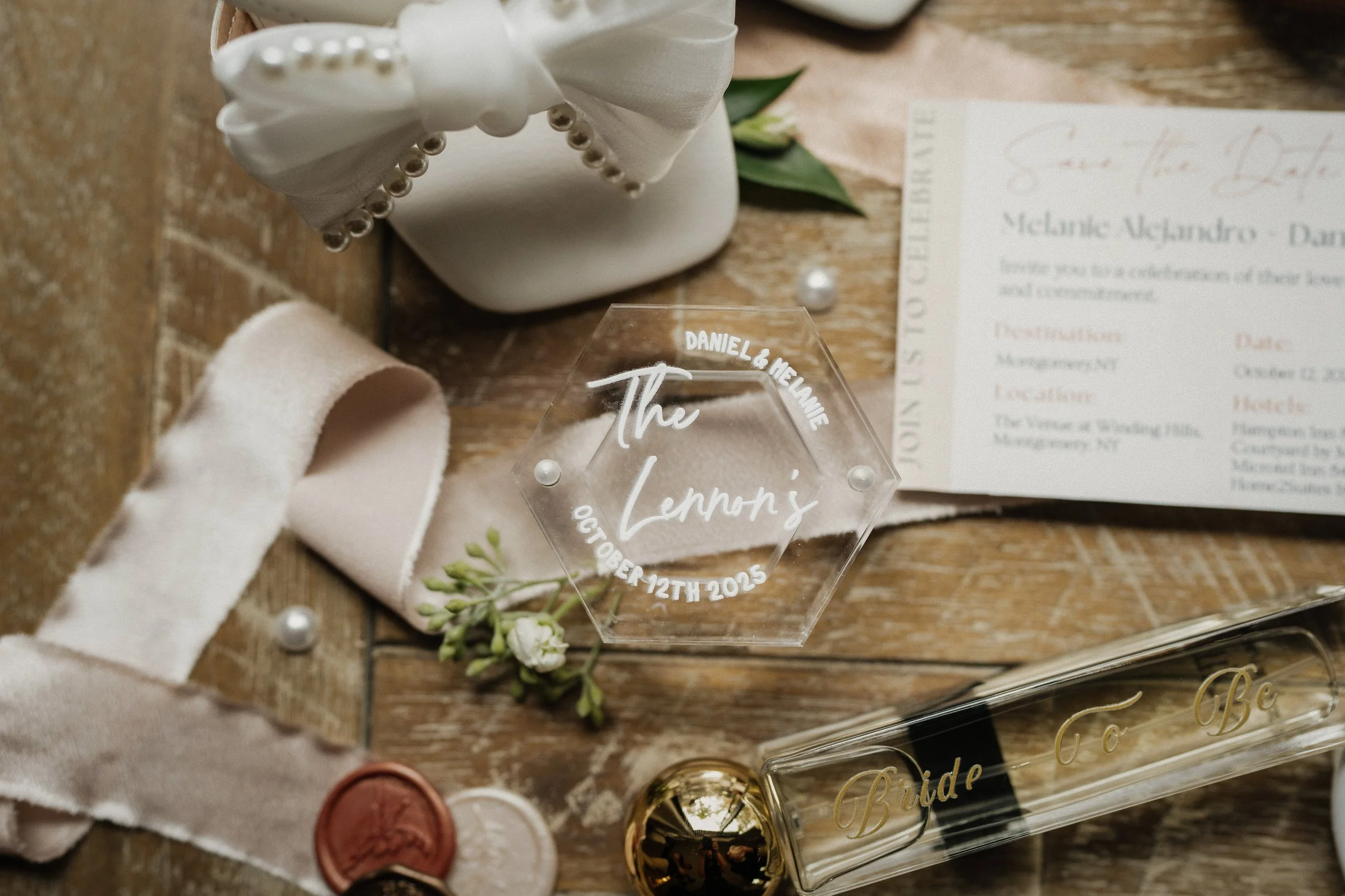 Wedding invitation, personalized hexagon acrylic keepsake with bride and groom's names and wedding date, boutonniere with small white flower, cream-colored sash with a wax seal, glass spray labeled 'Bride To Be,' partially visible wedding menu or pro