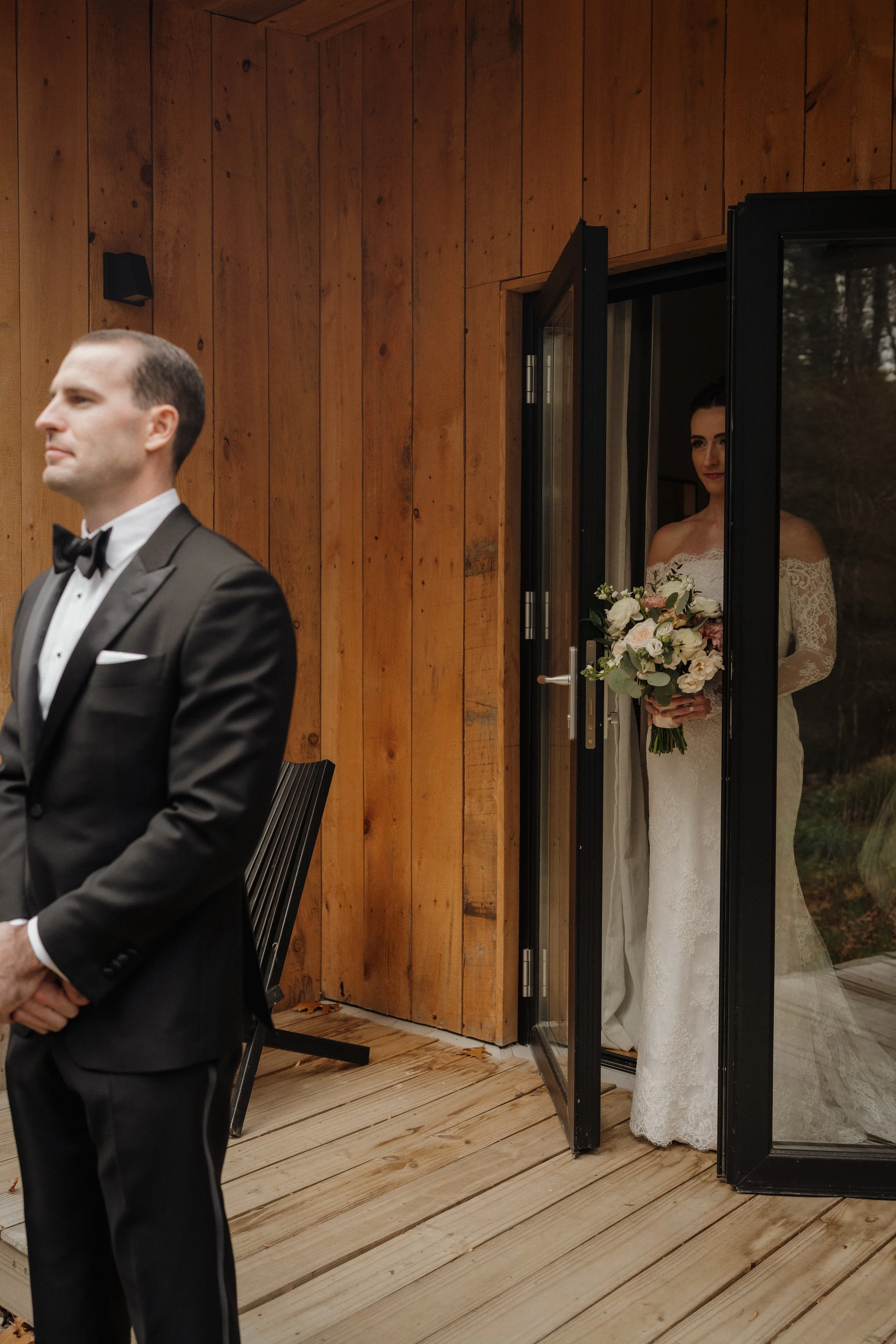 A bride in a white lace wedding gown holding a bouquet of flowers stands behind glass doors, observing a groom in a black tuxedo, who is standing in front of her with hands clasped.