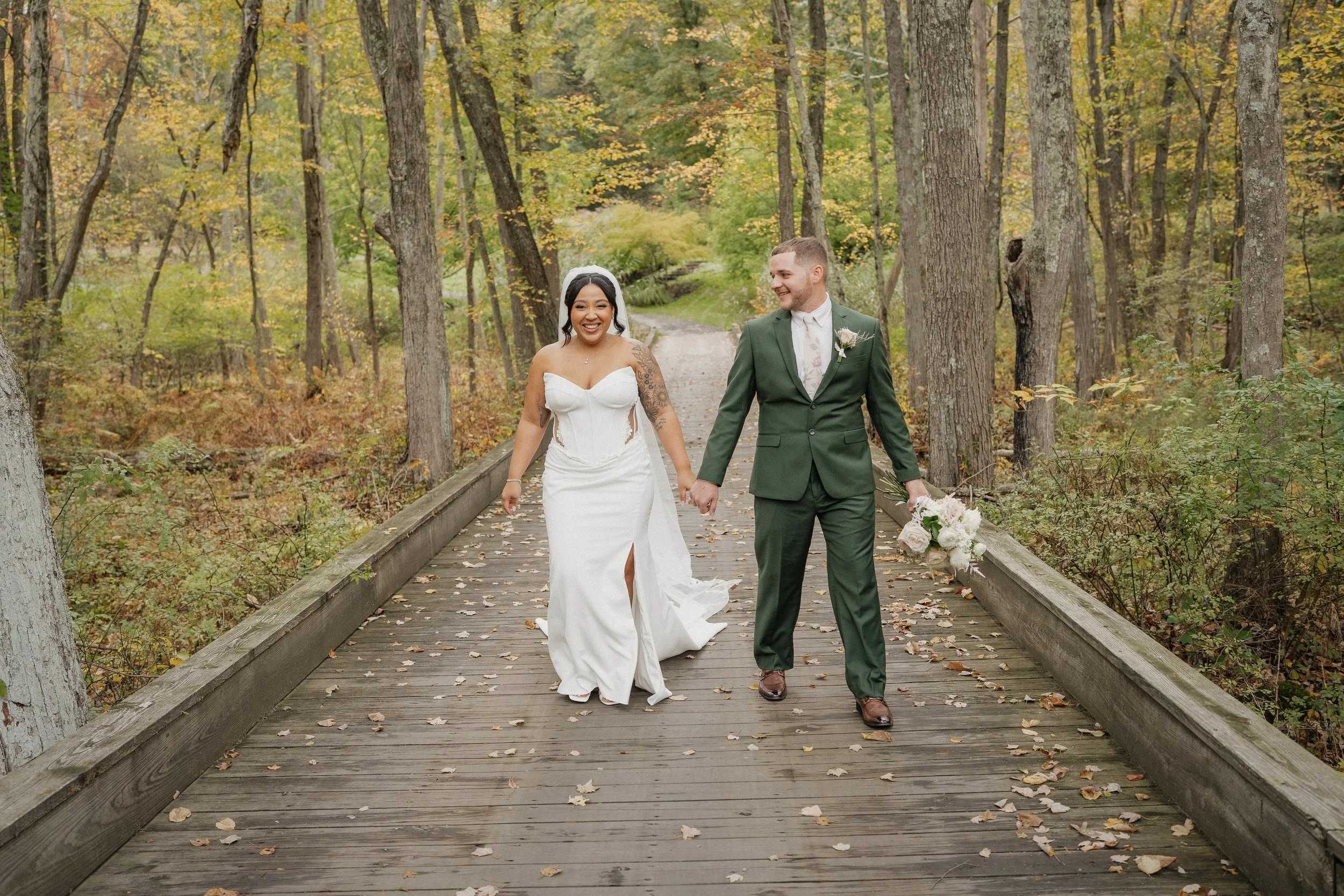 A newlywed couple walking hand-in-hand on a wooden bridge in a forest during autumn, with the bride in a white dress and the groom in a green suit holding a bouquet of flowers.