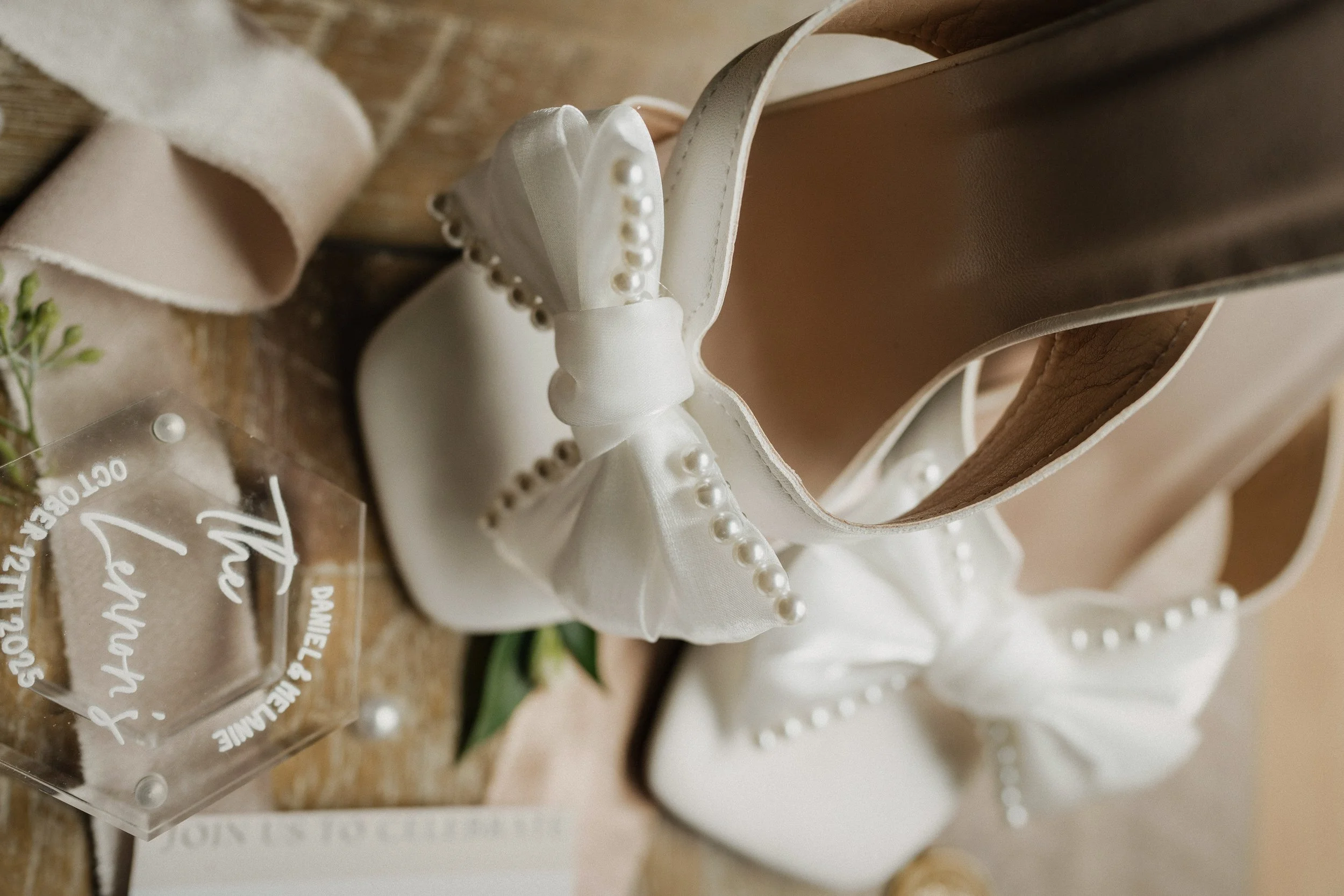 Close-up of a white high-heeled shoe with a large satin bow and pearl details, placed on a wooden surface with a beige ribbon and a small floral arrangement nearby.