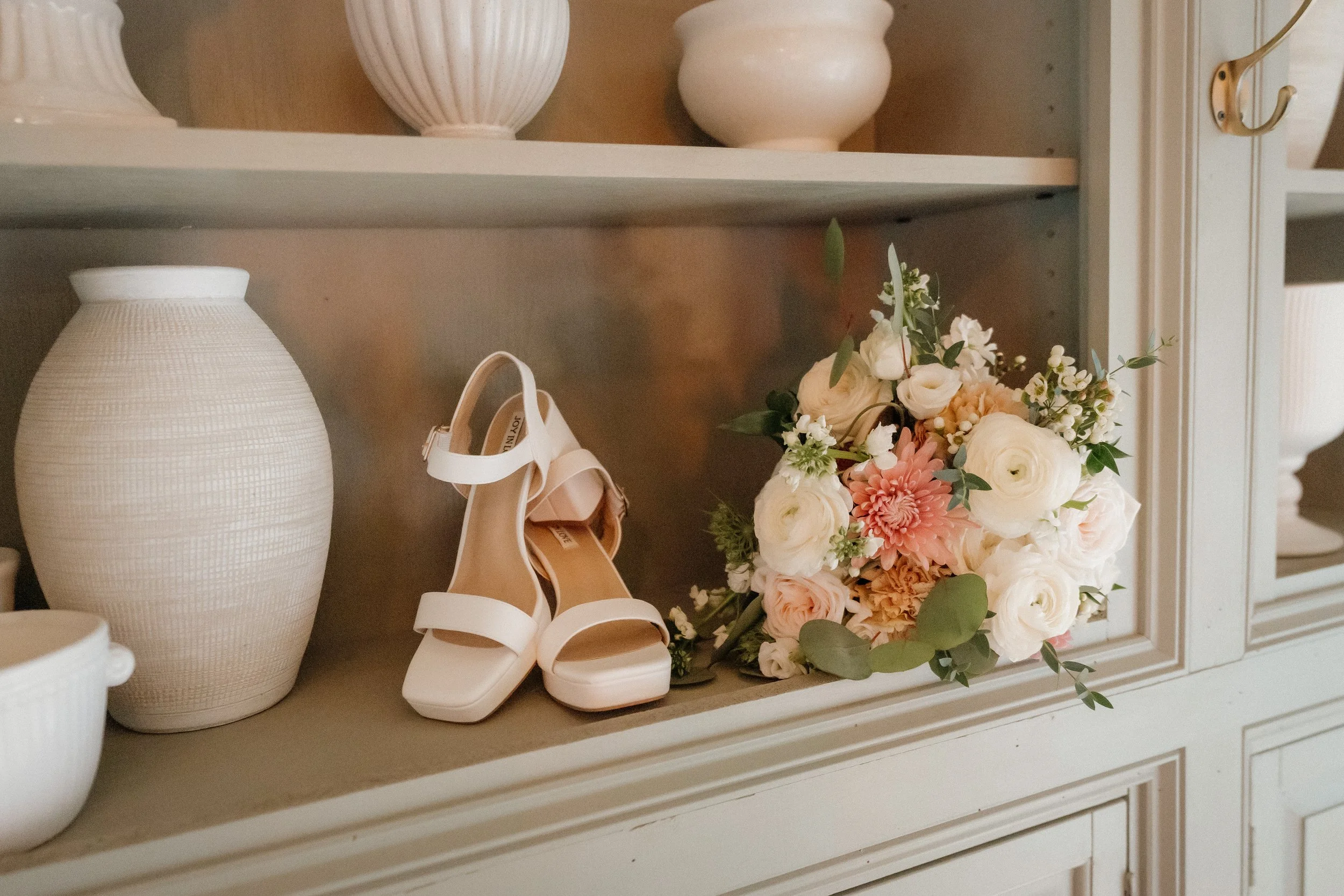 A white pair of high-heeled sandals with ankle straps placed on a beige shelf next to a bouquet of white and pink flowers and white ceramic vases.