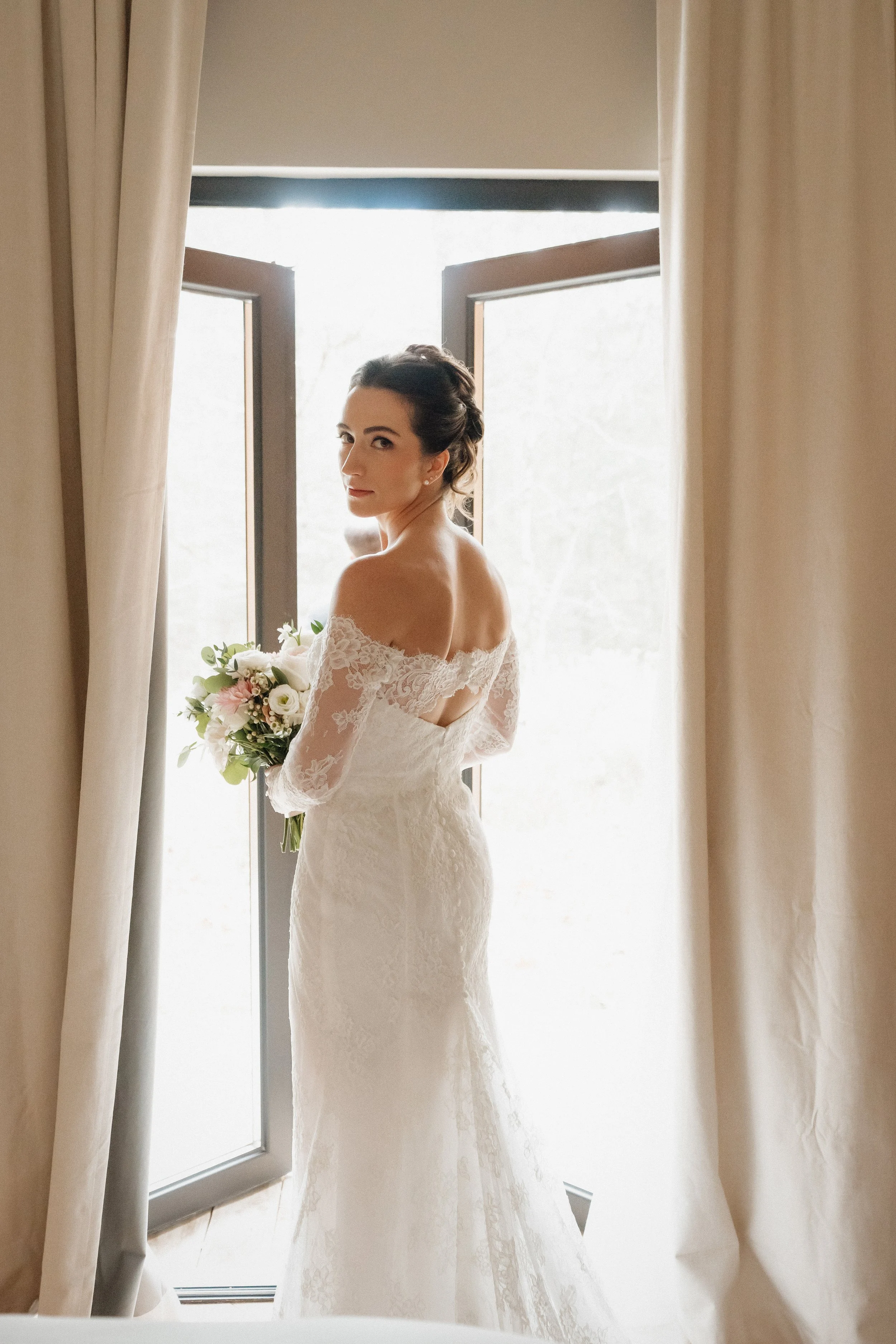 Bride in a white lace wedding gown holding a bouquet standing near open windows, looking back.