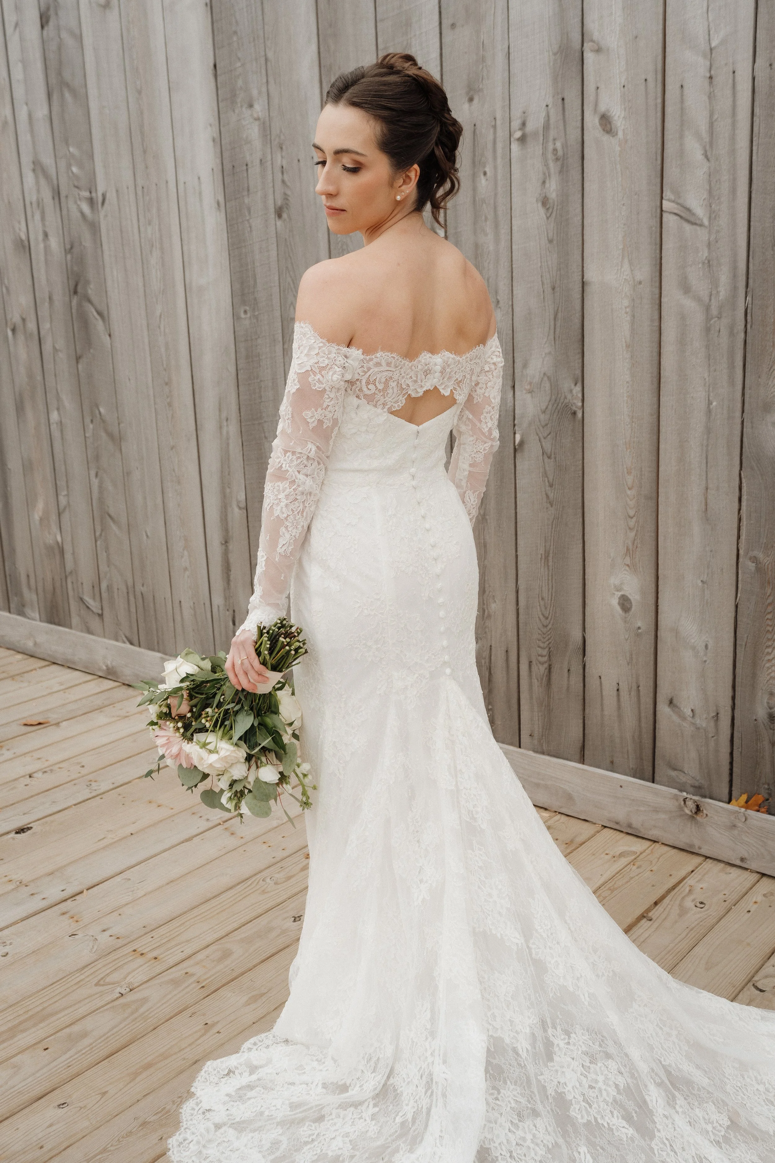 Bride in a white lace wedding gown holding a bouquet of pink and white flowers, standing on a wooden deck with a wooden fence in the background.