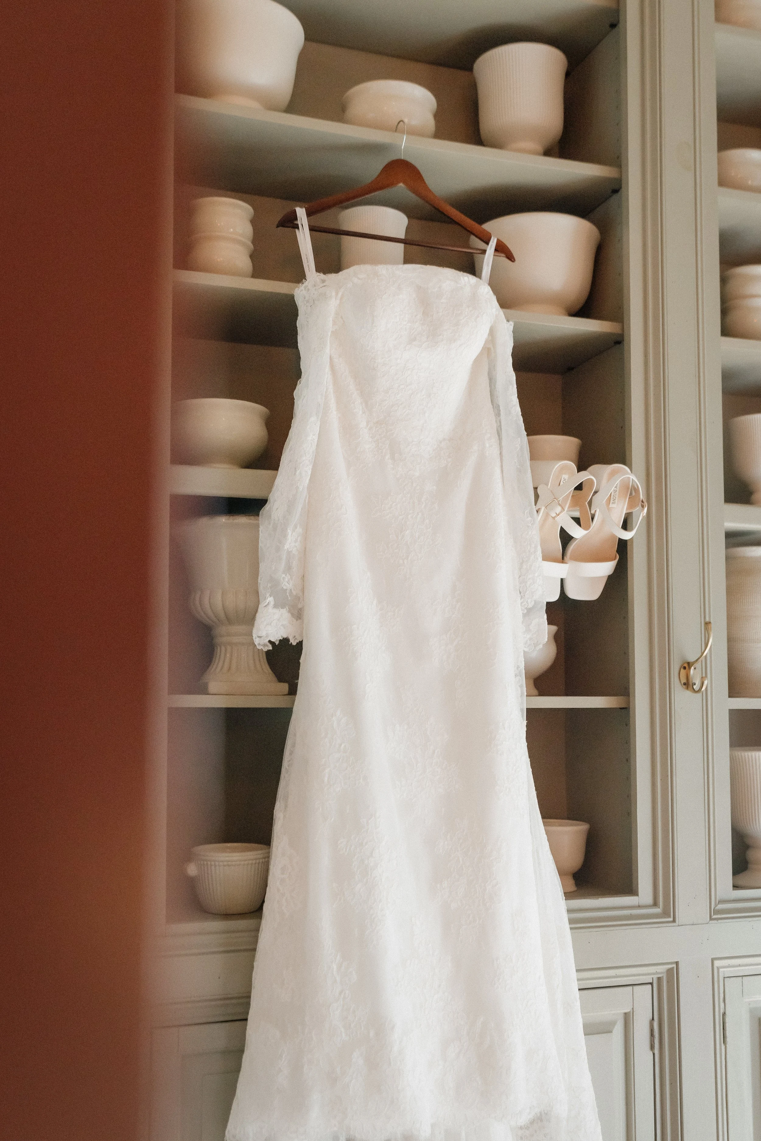 A white wedding dress hanging on a wooden hanger inside a cabinet with white ceramics and bowls on the shelves.
