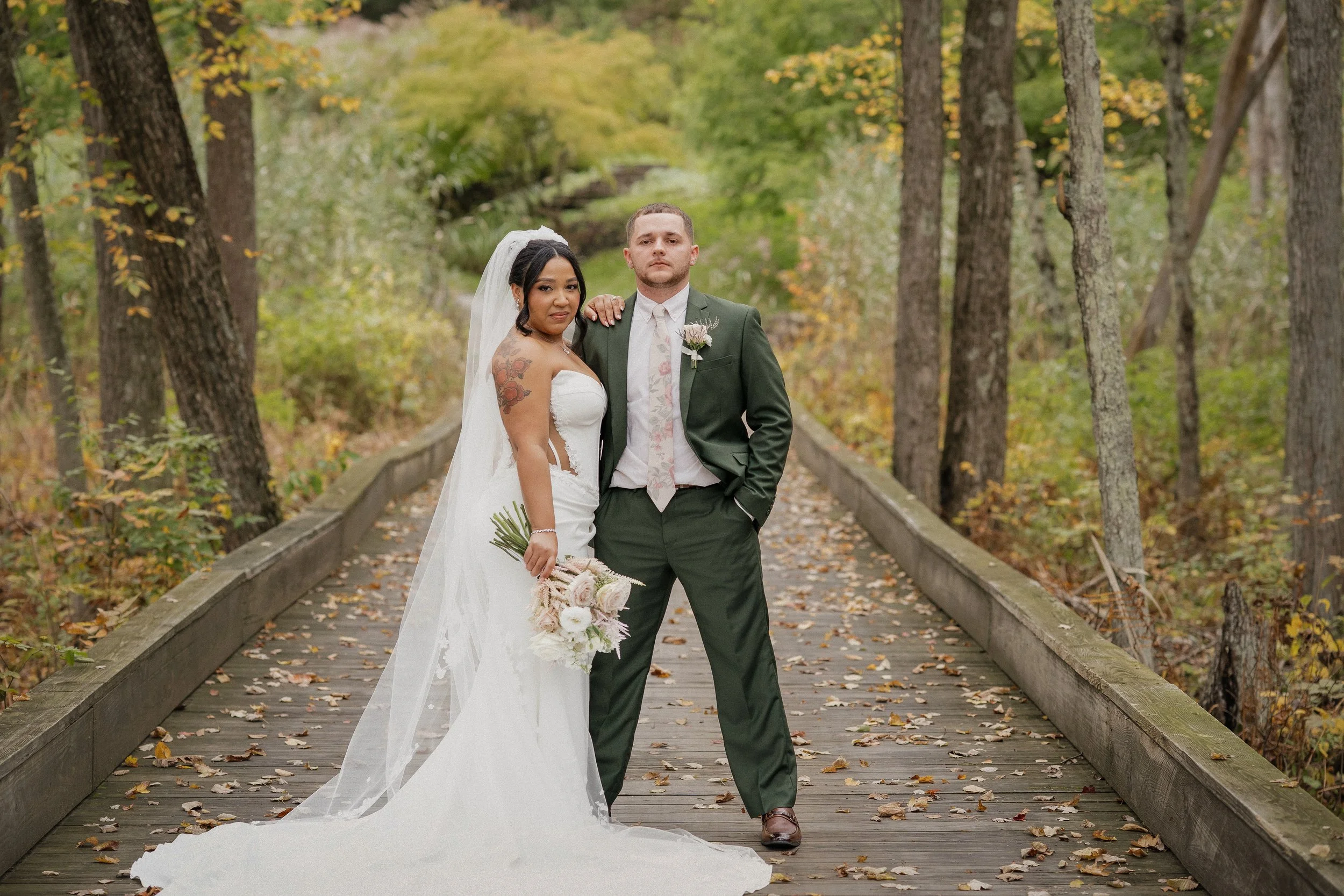 Bride and groom standing together on a wooden bridge in a wooded area, with fall foliage in the background, during their wedding.