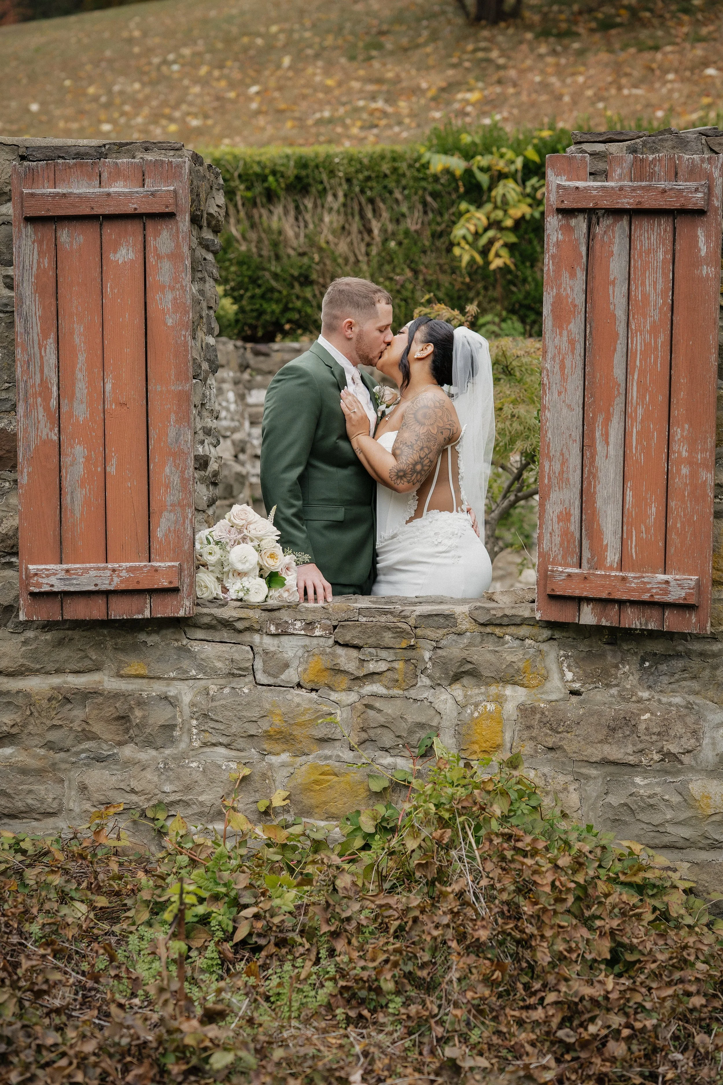 A newlywed couple sharing a kiss in a stone window with red shutters, outdoors surrounded by autumn foliage.