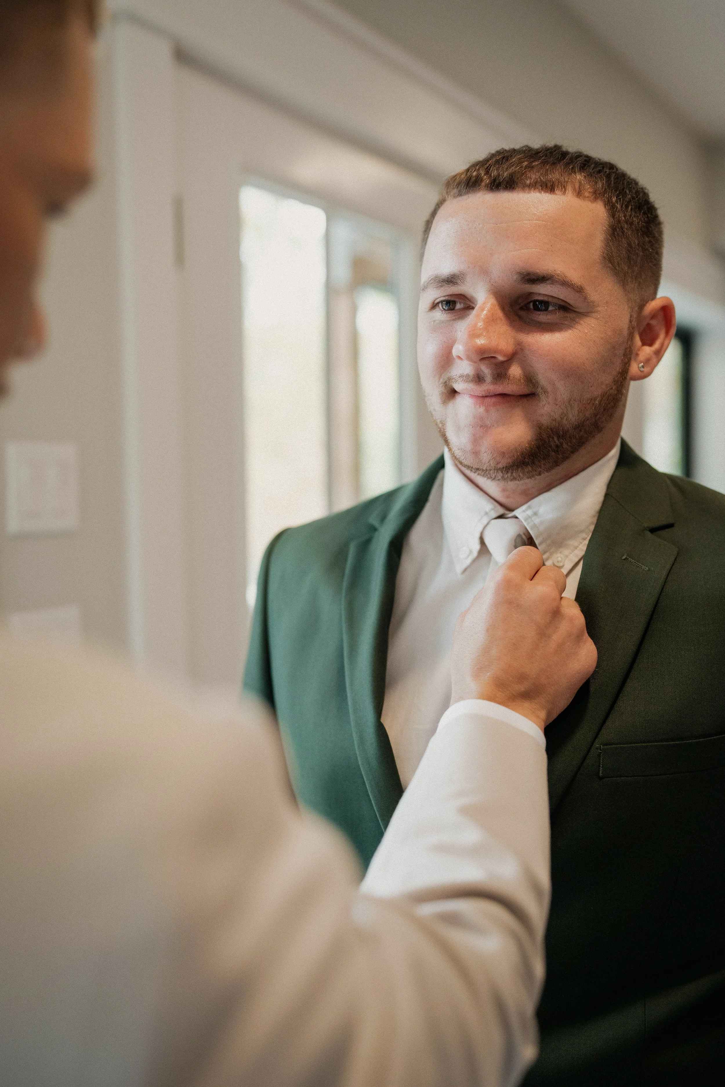 A man in a green suit jacket and white shirt is being helped to adjust his tie by another person whose arm is visible in the foreground.