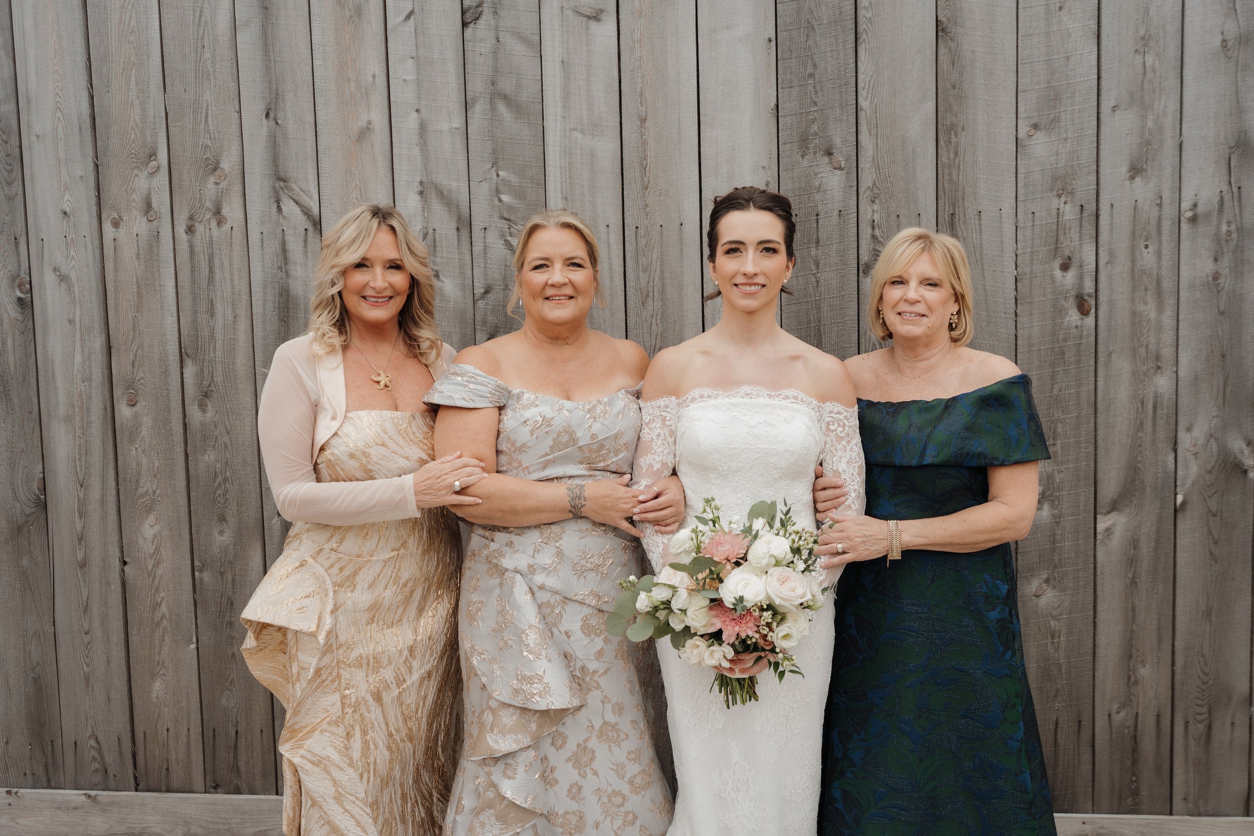 Four women standing together outdoors against a wooden fence, with the bride in the center holding a bouquet of flowers, all dressed in formal attire.