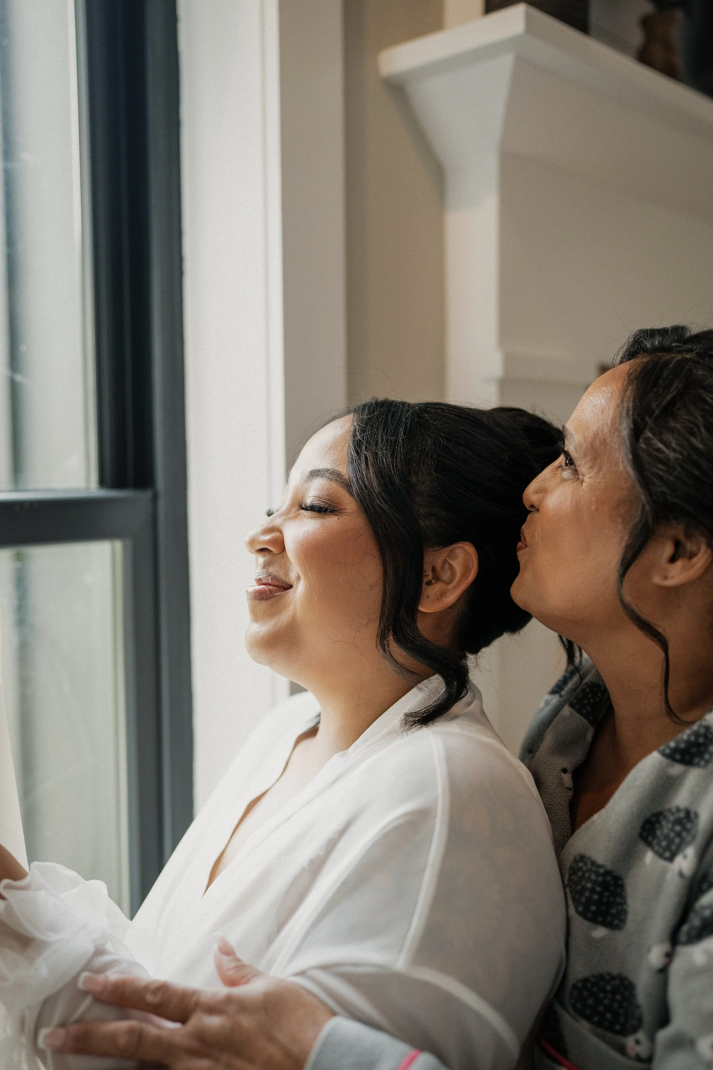 A woman smiling happily as she looks out the window, with another woman leaning close to her, possibly providing support or sharing a moment of joy.