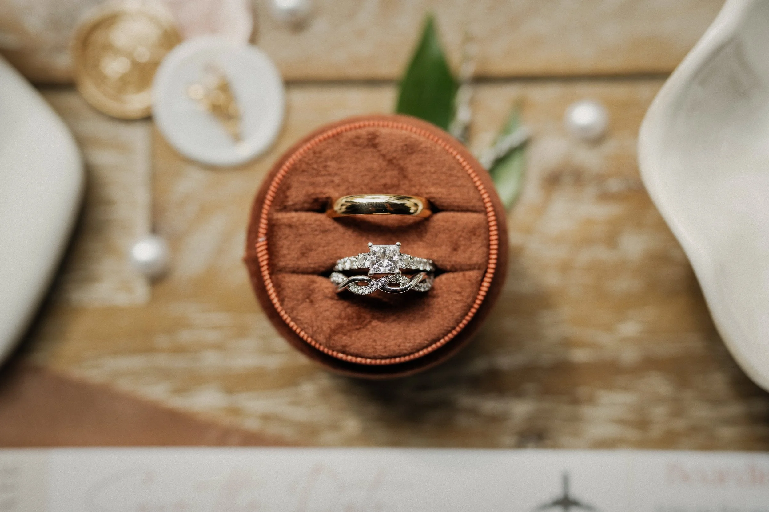 Close-up photo of a round brown jewelry box with two rings inside on a wooden surface, surrounded by small decorative items and pearls.