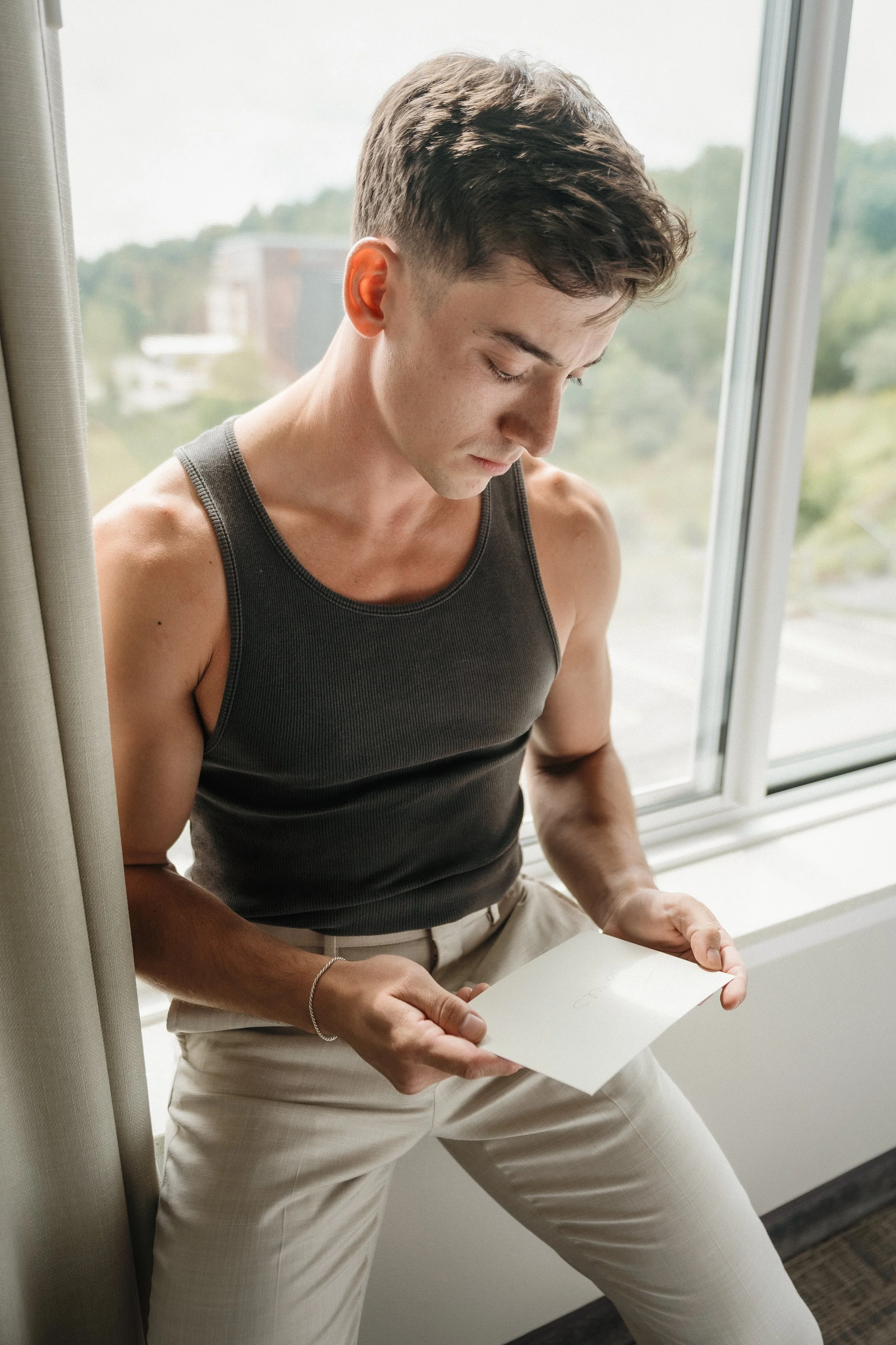A young man with dark hair in a black sleeveless shirt and light pants sitting by a window, looking down at a white envelope in his hands.