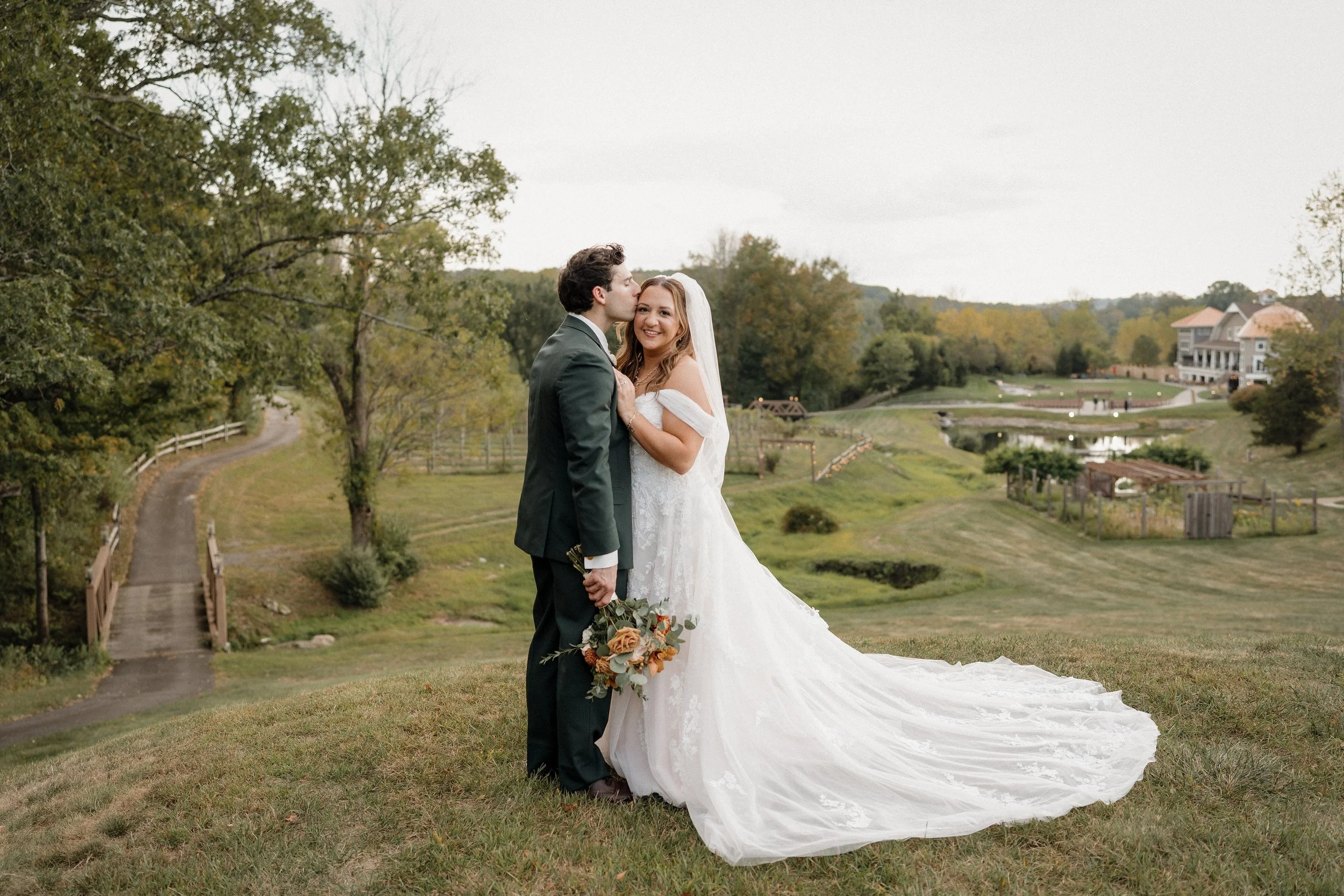 A bride and groom standing together outdoors on a grassy hill, with the groom kissing the bride on her temple. The bride is holding a bouquet of flowers and is smiling. They are dressed in wedding attire, with the bride in a white lace gown and the groom in a black suit. In the background, there is a winding path, trees, and a large house or estate on a hill.