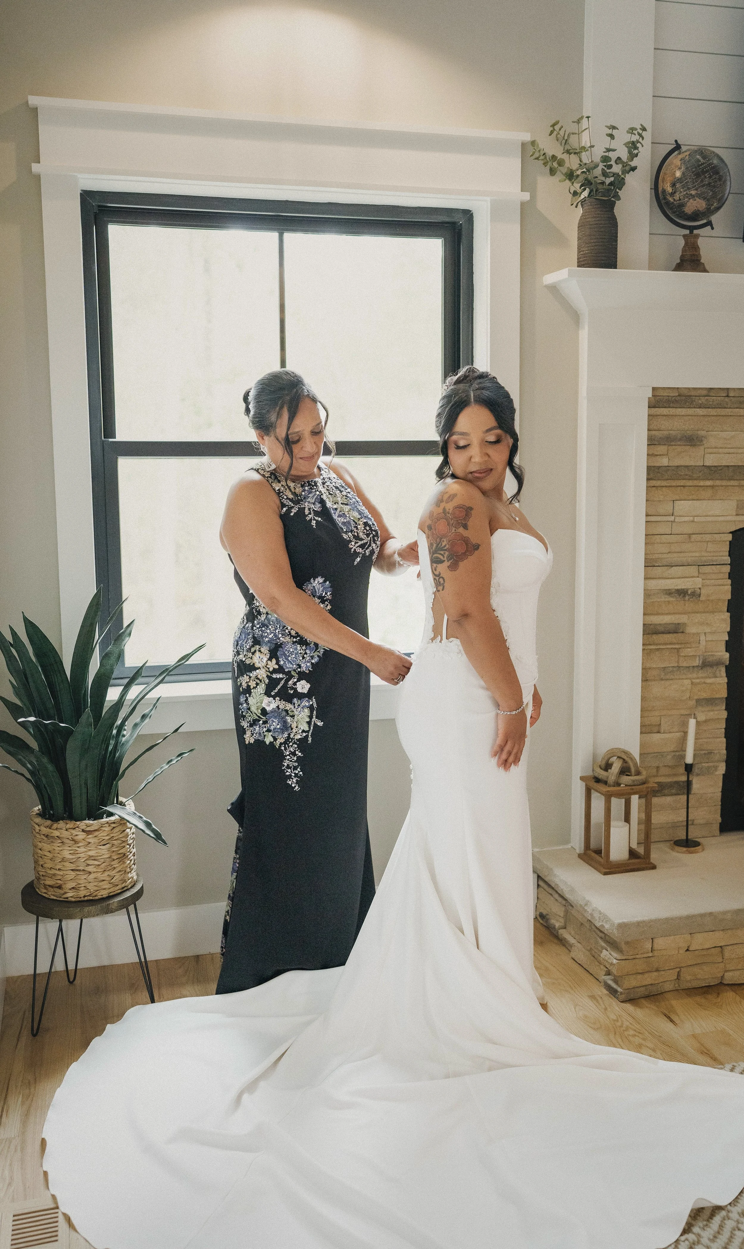 A bride in a strapless white wedding dress with tattoos on her arm stands in a room, as another woman helps her prepare, possibly for her wedding.