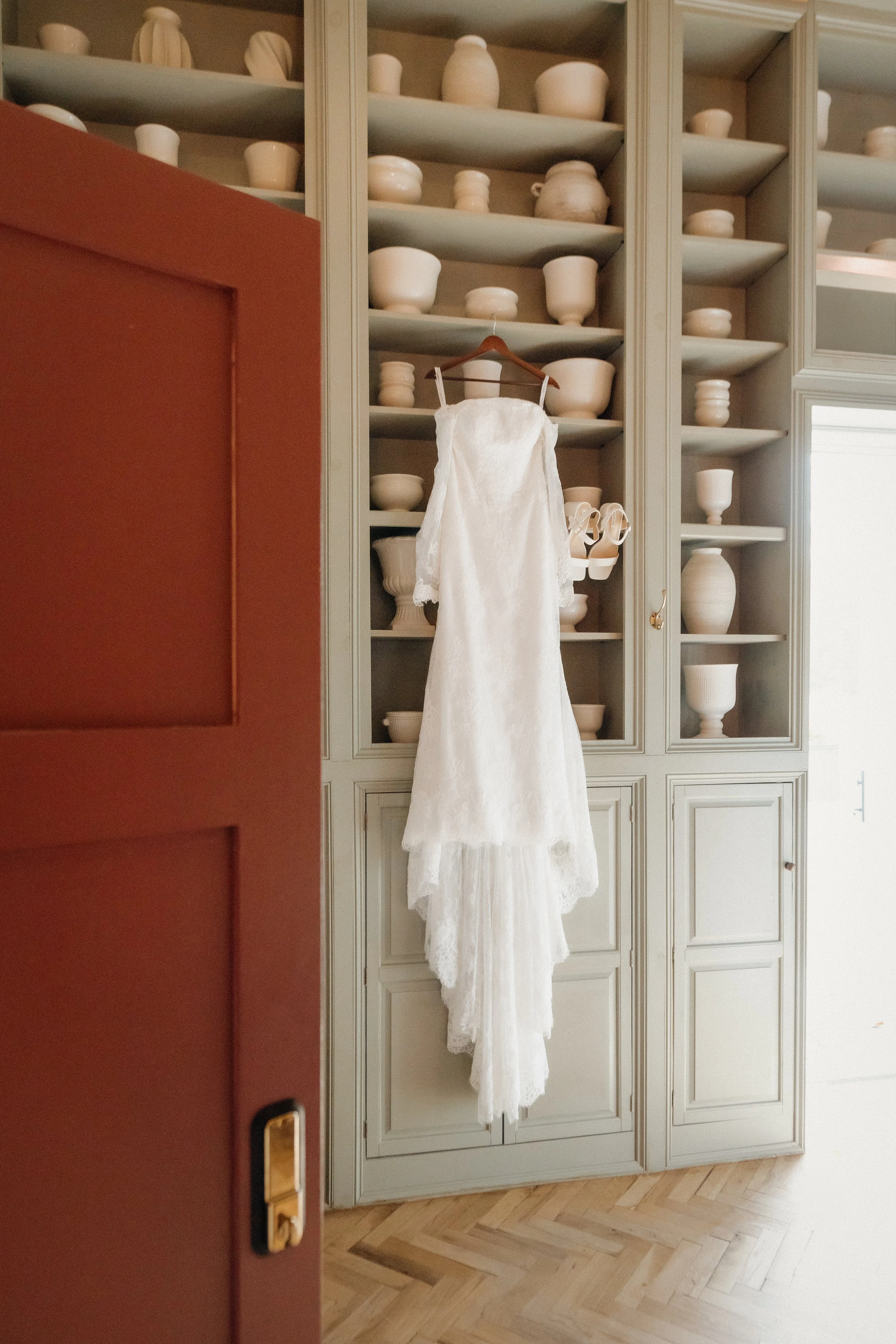 A white wedding dress hanging on a wooden hanger on a gray cabinet with open shelves filled with white pottery.