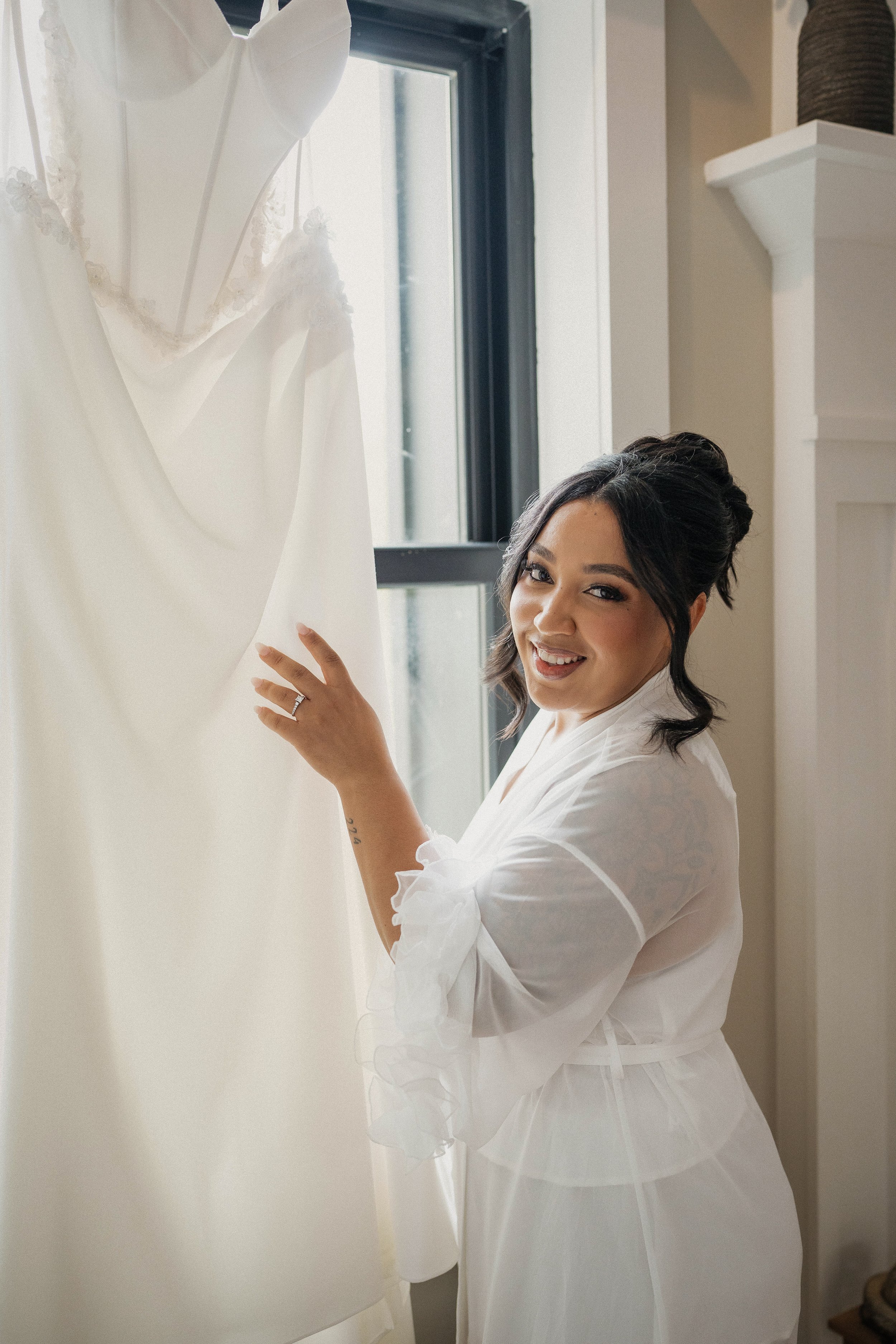 A woman with dark hair styled in an updo, wearing a white robe, smiling while holding a white wedding dress near a window.