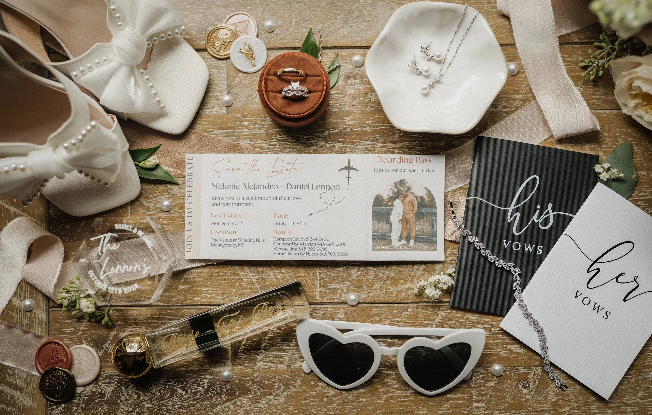 Wedding invitation with jewelry, shoes, and accessories arranged on a wooden table. The invitation is for Melanie Alejandro and Daniel Lennon, scheduled for October 12, 2025, in Montgomery, NY. Items include white shoes with bows, pearl jewelry, a vo