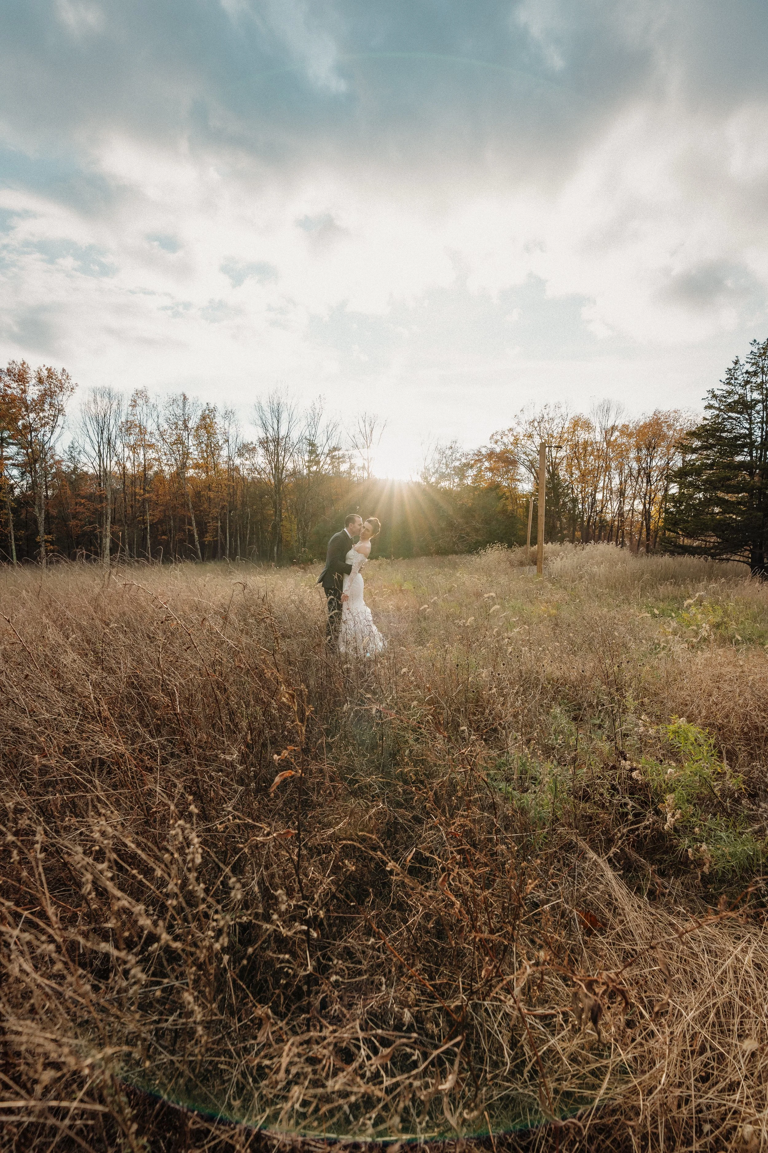A bride and groom dancing in a field at sunset, with trees in the background and a partly cloudy sky.