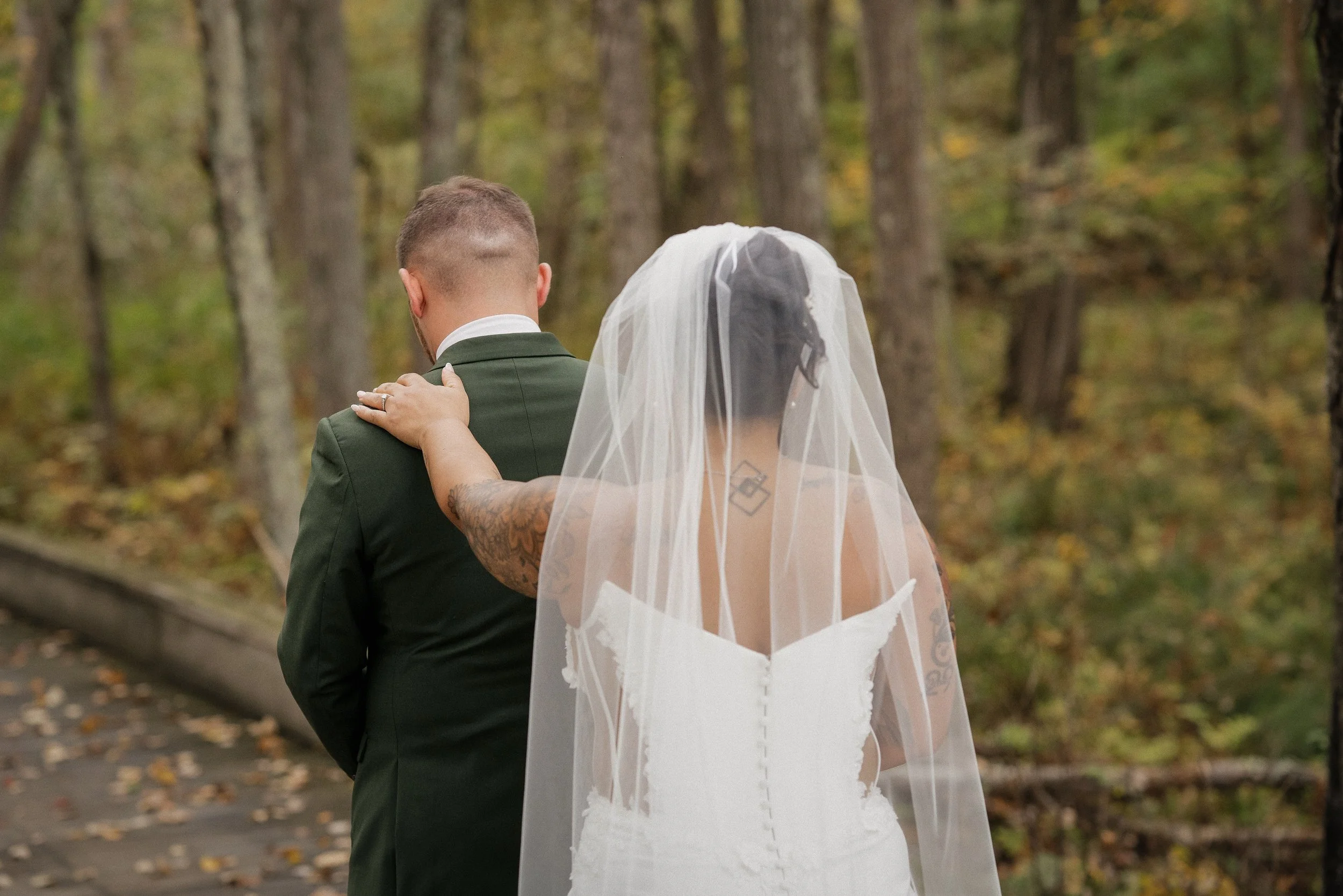 A bride and groom standing in a wooded area during their wedding, with their backs to the camera. The bride has tattoos on her arms and wears a white wedding gown with a veil, while the groom is dressed in a dark green suit.