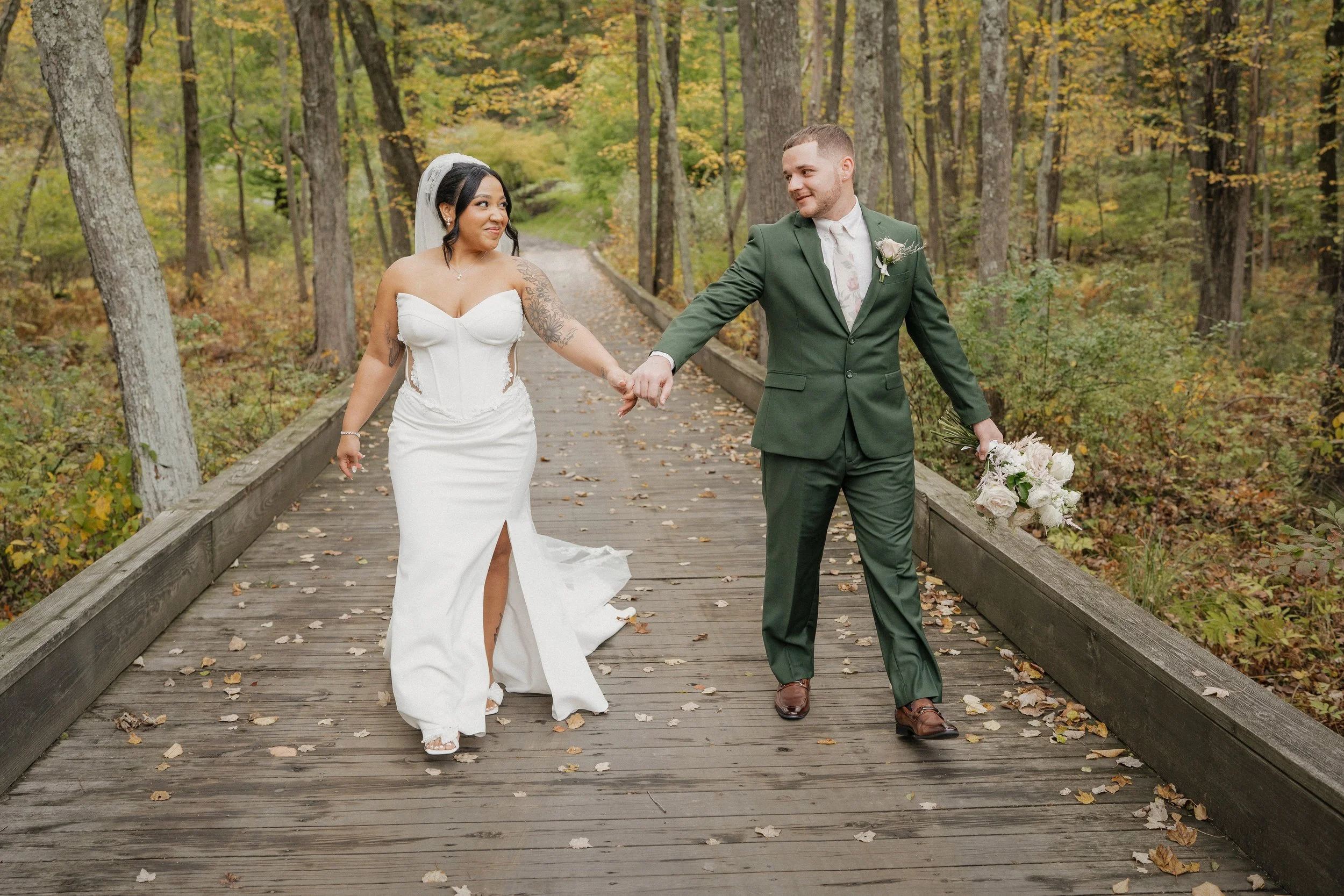 A bride and groom walking hand in hand on a wooden bridge in a wooded area during autumn, with trees displaying fall foliage.