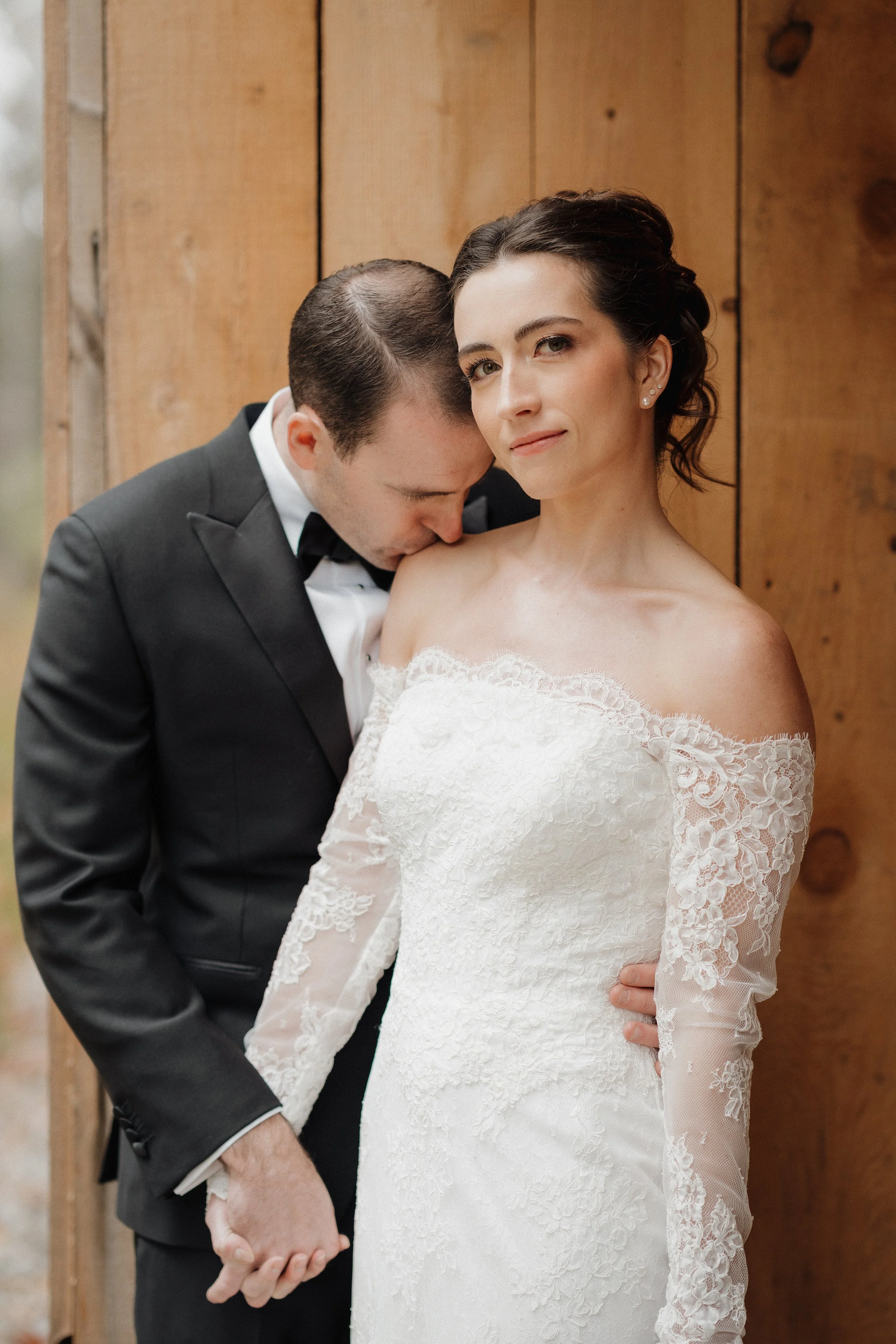 Bride and groom on their wedding day, with the groom kissing the bride's shoulder as she looks at the camera, wearing a white lace off-the-shoulder wedding dress against a wooden background.