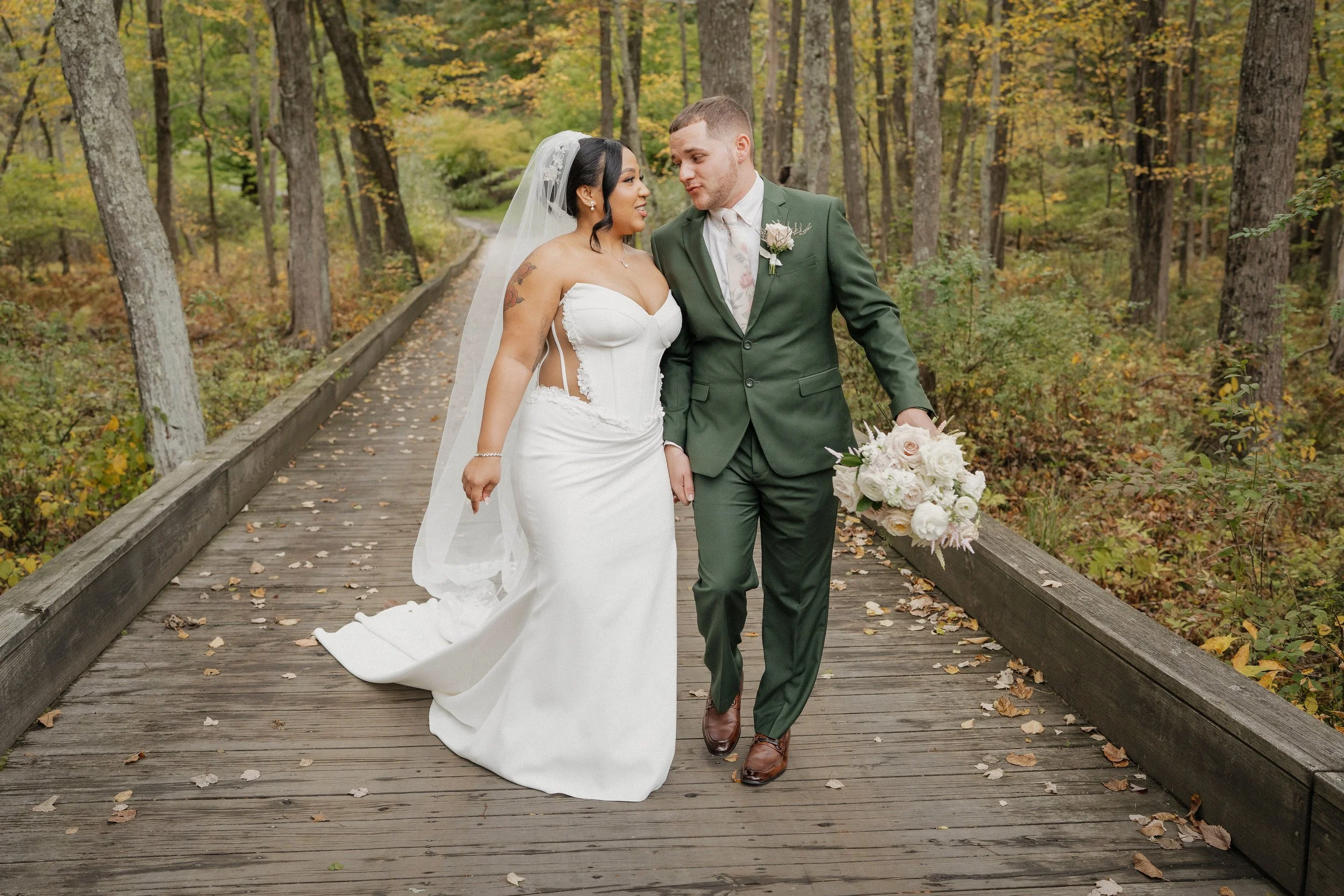 A newlywed couple walking on a wooden bridge in a forest during fall, with the bride in a white wedding gown and the groom in a dark green suit, holding a bouquet of white flowers.