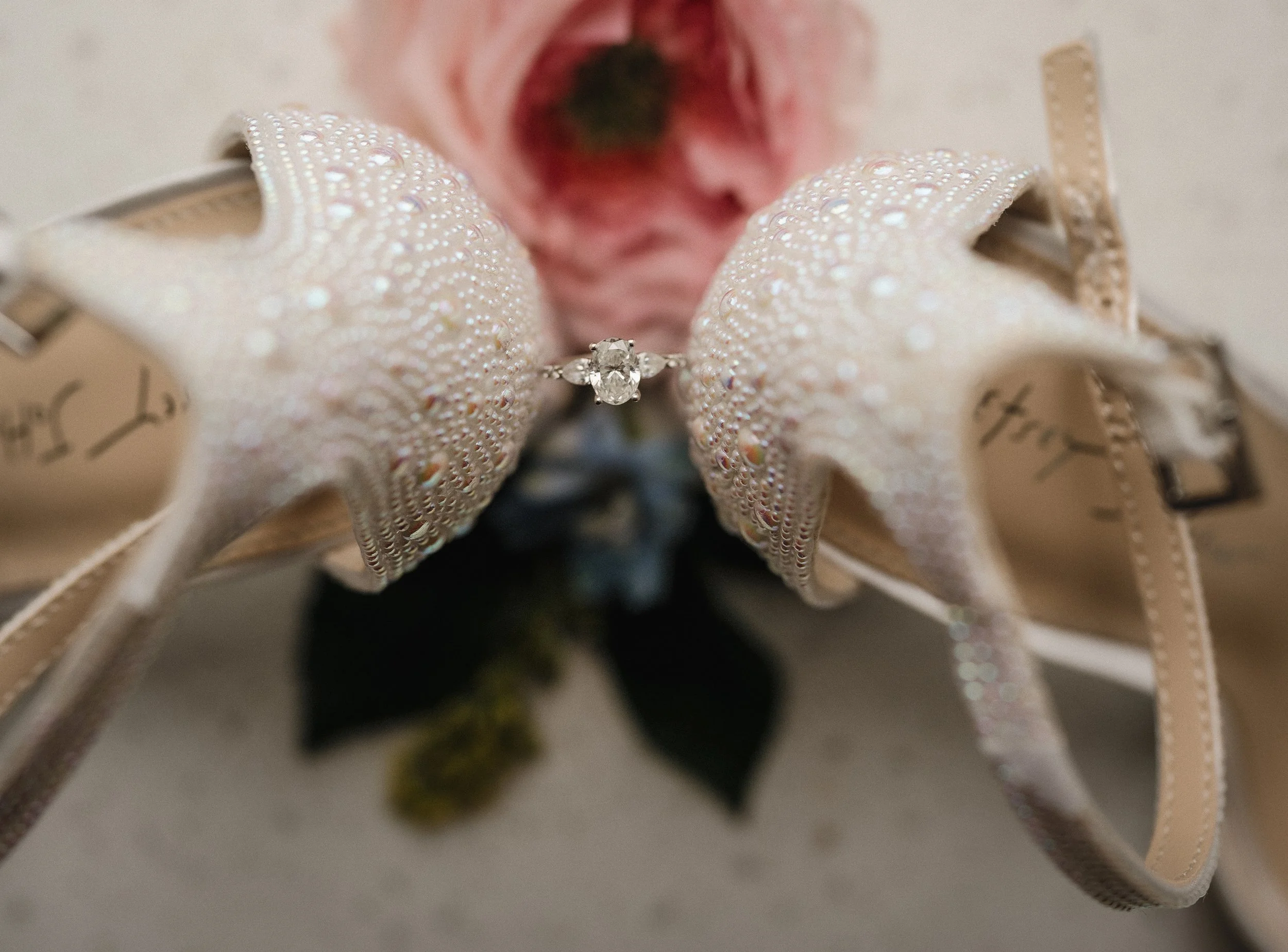 Close-up of a silver engagement ring with a central diamond, placed on a decorative starfish with shiny embellishments, accompanied by a pink rose and beige sandals with straps in the background.