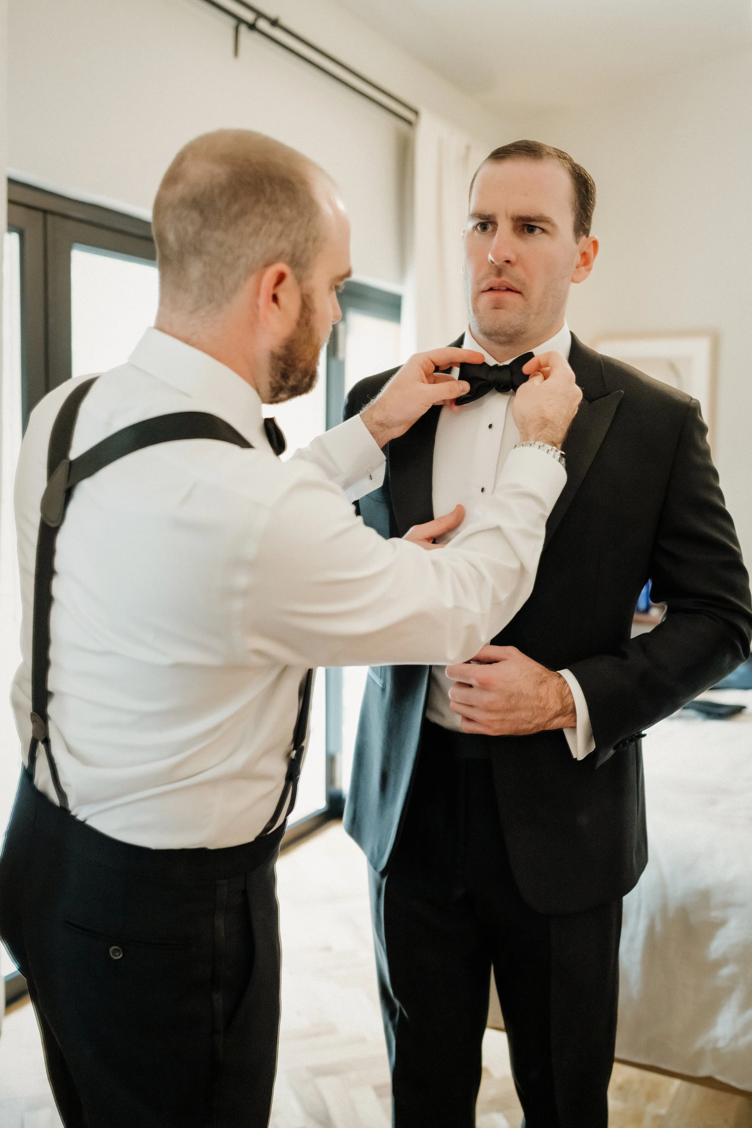 Two men in tuxedos, one helping the other adjust a bow tie in a room with natural light.