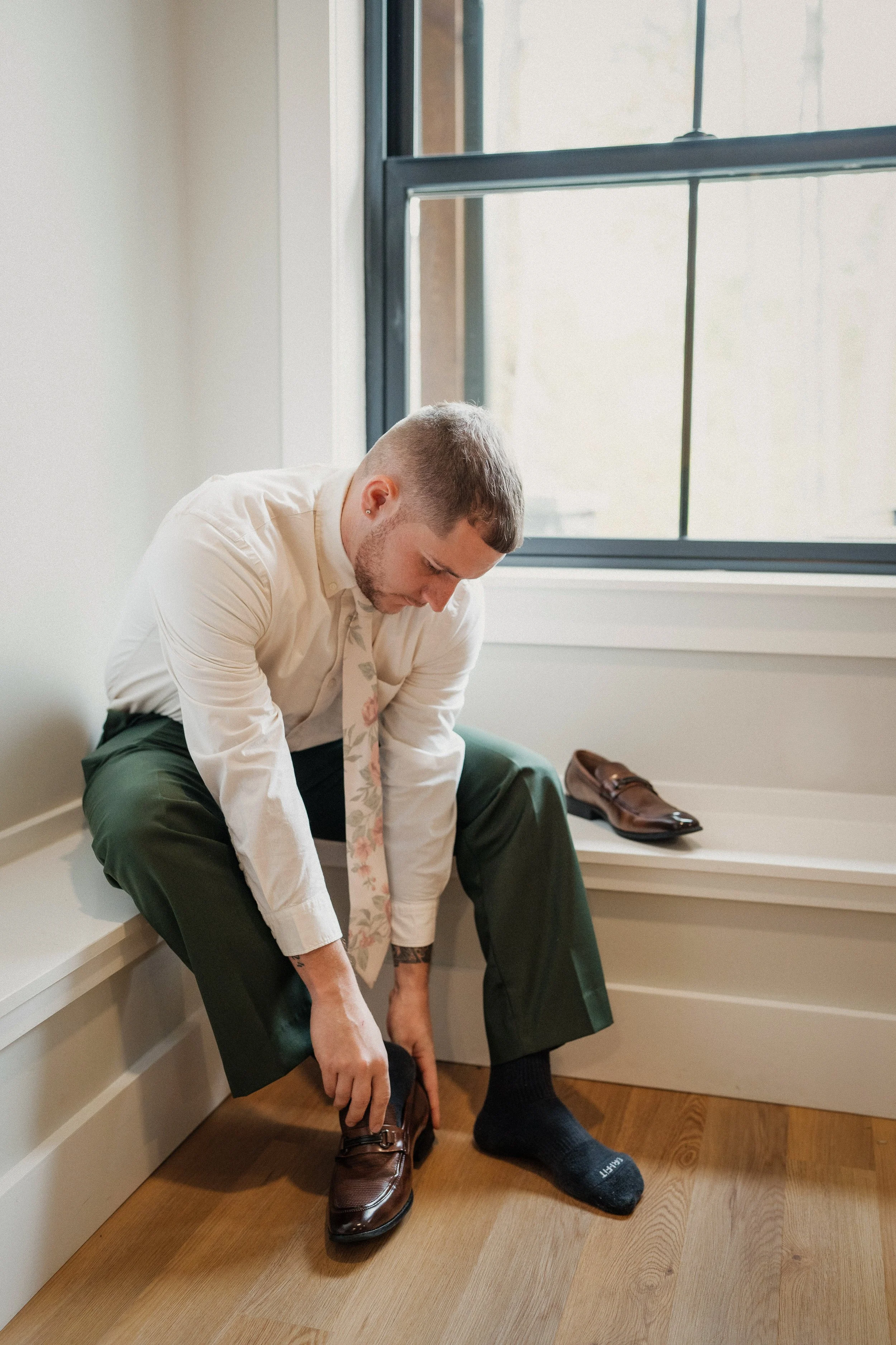 A man dressed in formal attire, including a white shirt, floral tie, green pants, and dark socks, is sitting on a bench near a window, putting on brown leather shoes.