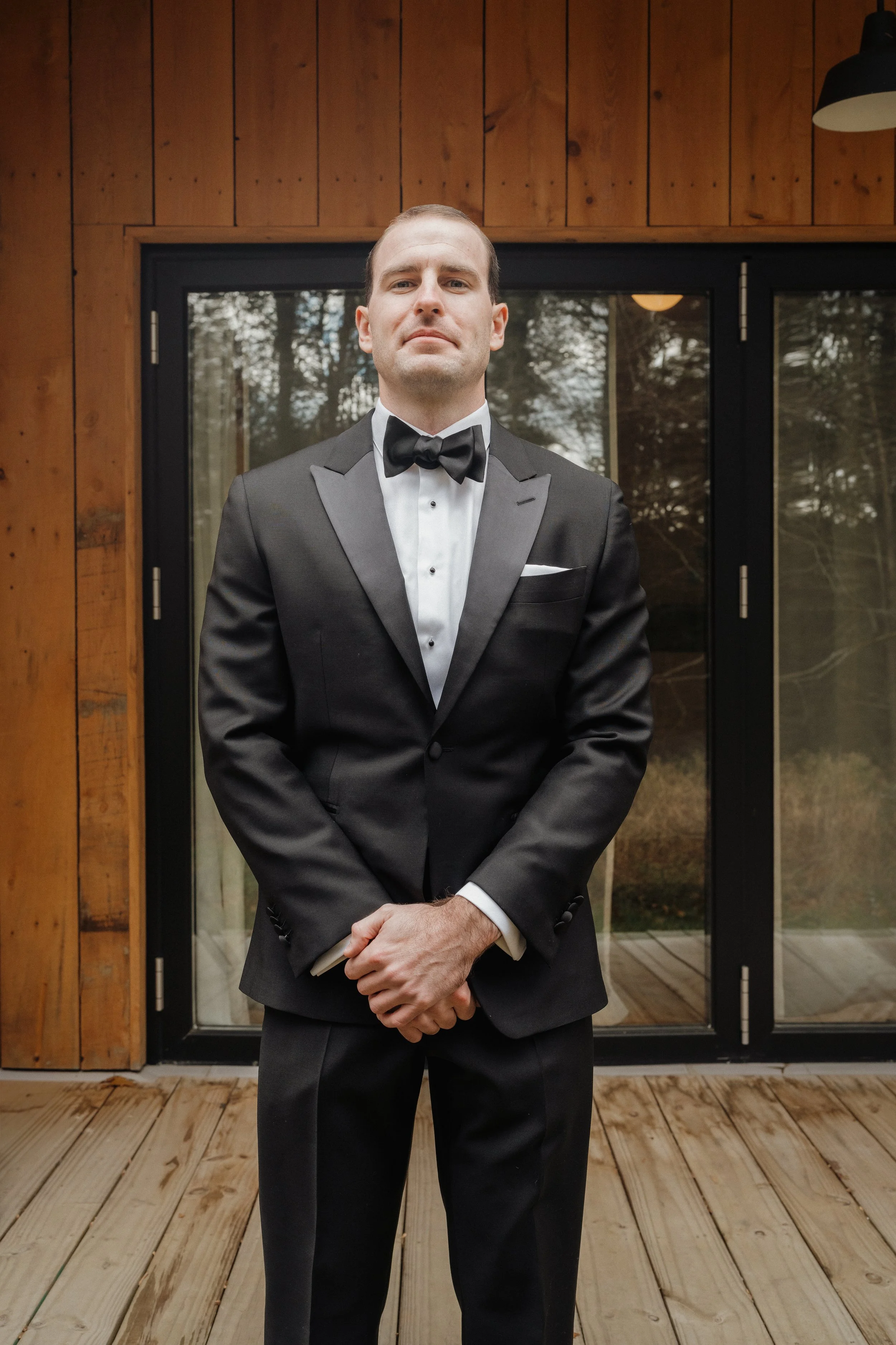 A man in a black tuxedo, white dress shirt, and black bow tie standing on a wooden deck in front of glass doors with a reflection of trees outside.