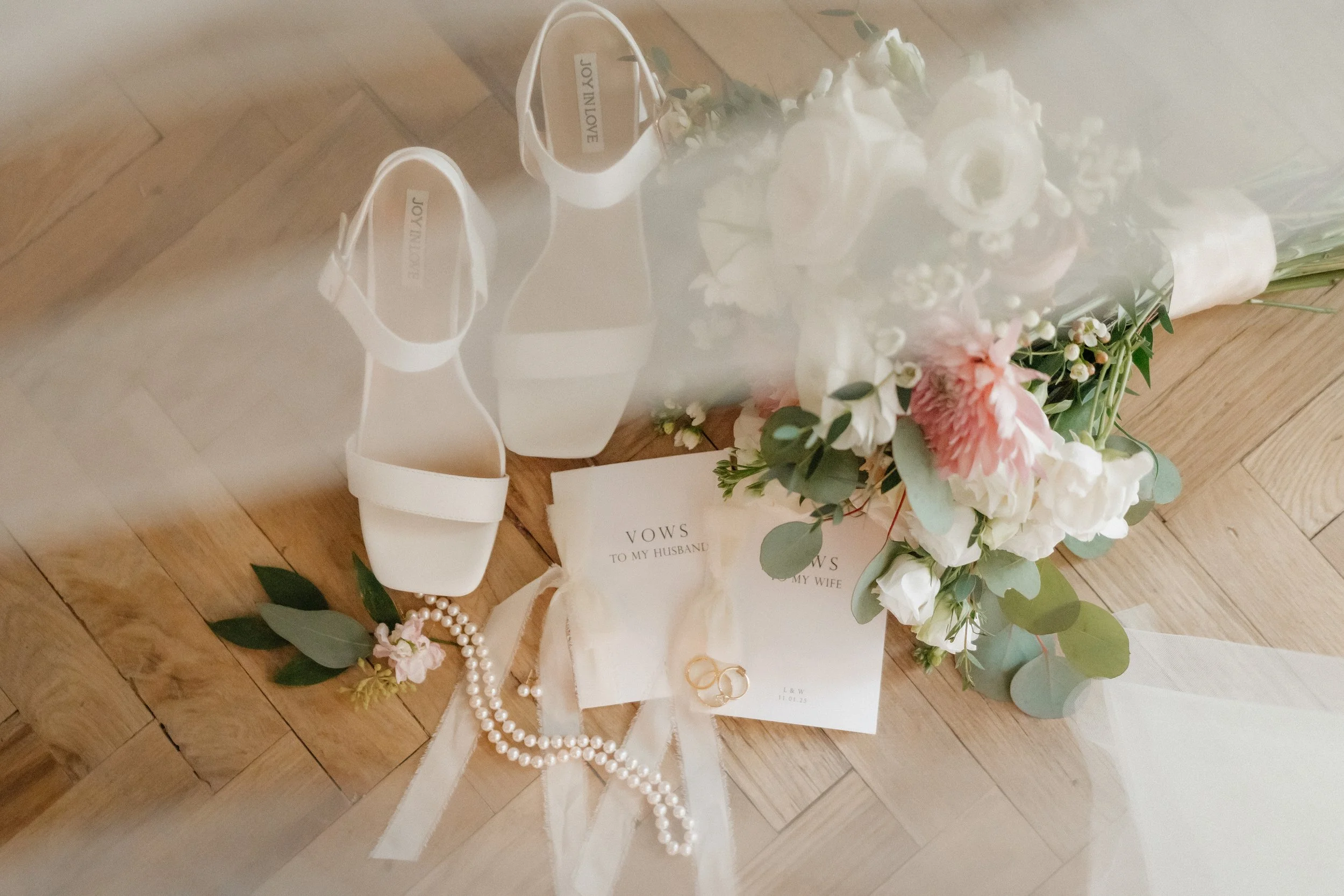 Wedding bouquet with pink and white flowers, white high-heeled shoes, wedding rings, vow notes, and pearl necklace on wooden floor.