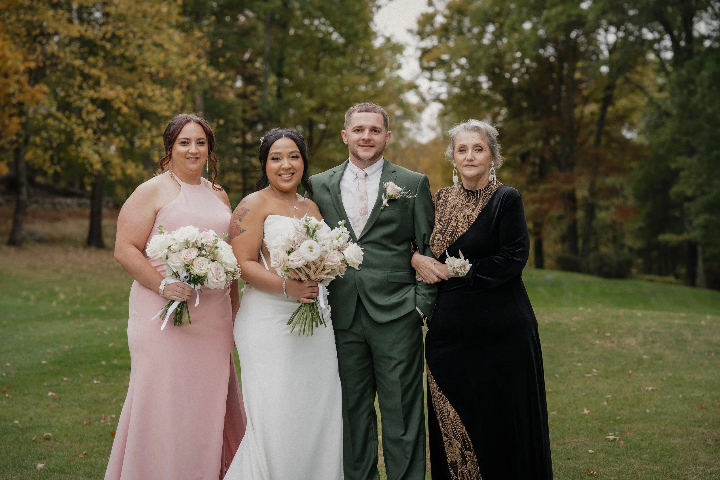 A wedding party outdoors with four people: two women in dresses, one man in a green suit, and one older woman in black and gold attire, standing on grass with trees and autumn foliage in the background.