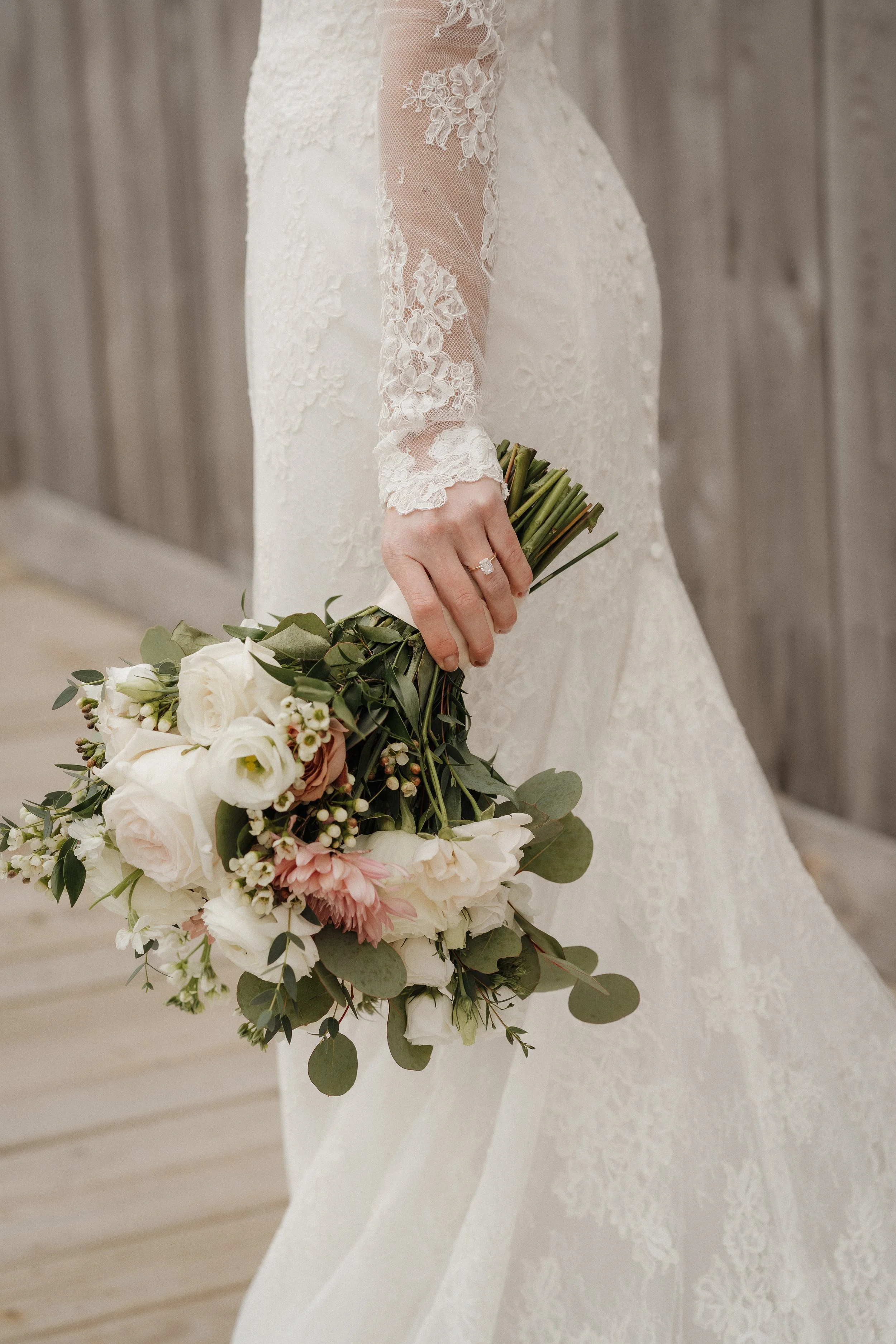 A bride holding a bouquet of white and blush pink flowers with greenery, wearing a lace wedding dress with long sleeves.