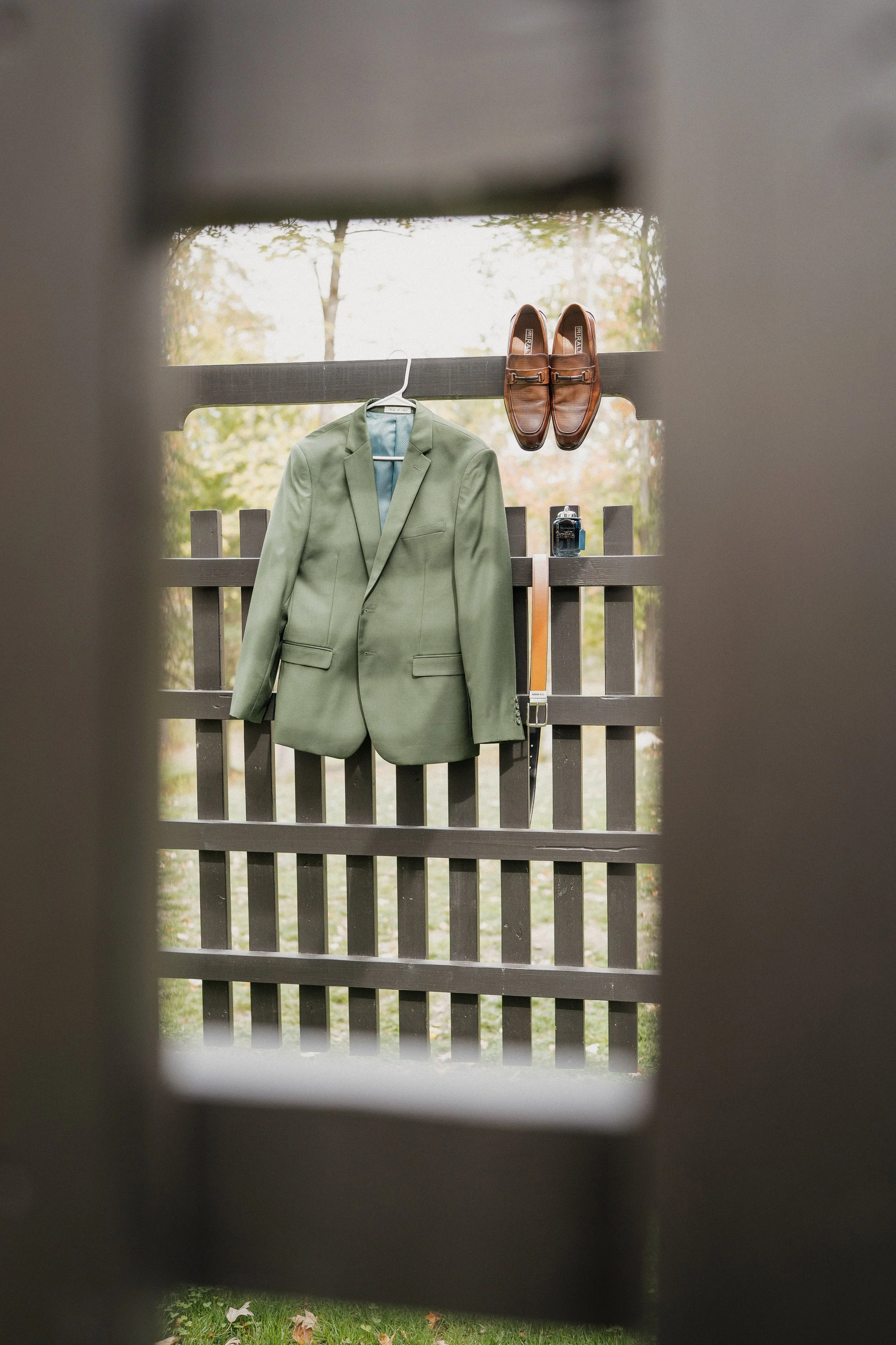 A green blazer hangs on a hanger on a wooden fence, with a pair of brown leather loafers and a small camera placed on the fence.